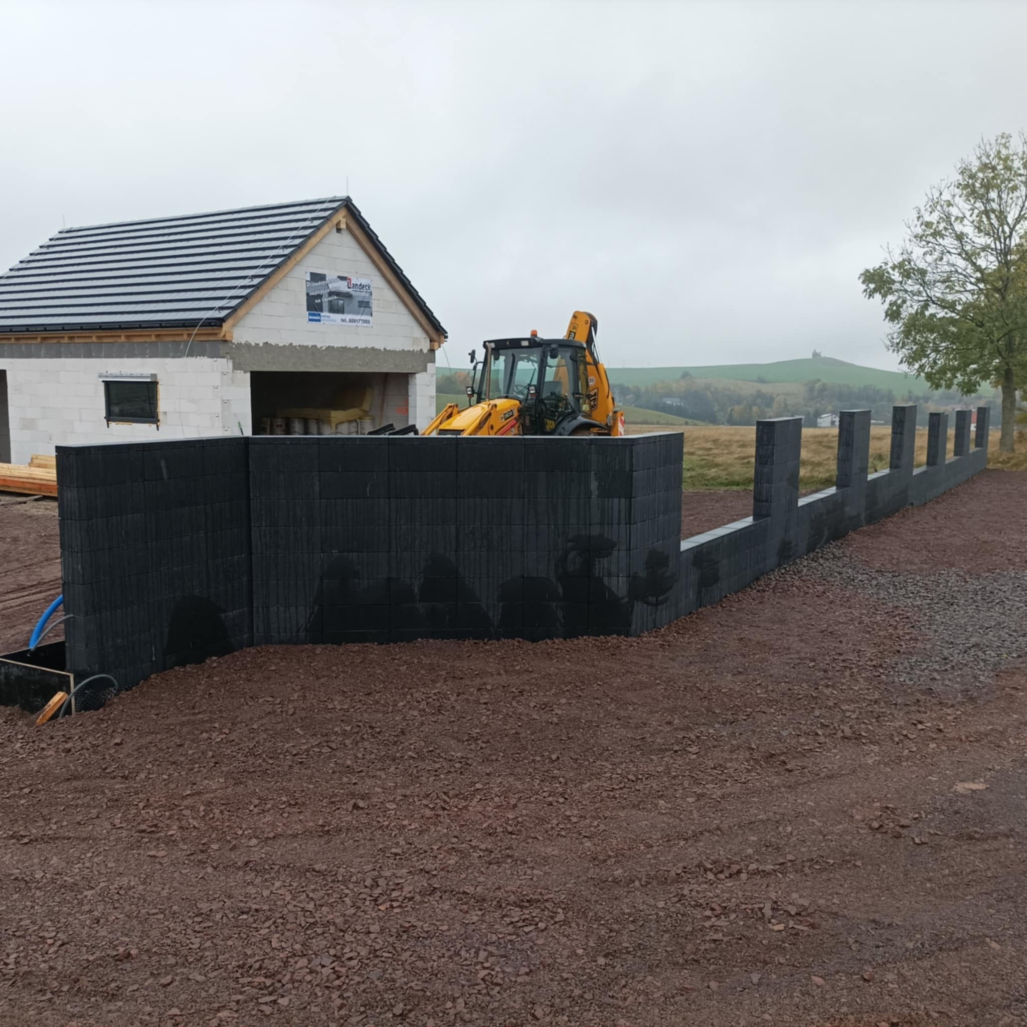 Częściowo wzniesione ogrodzenie betonowe z pustaków w kolorze grafitowym, z żółtą koparką w tle i budynkiem w stanie surowym otwartym.