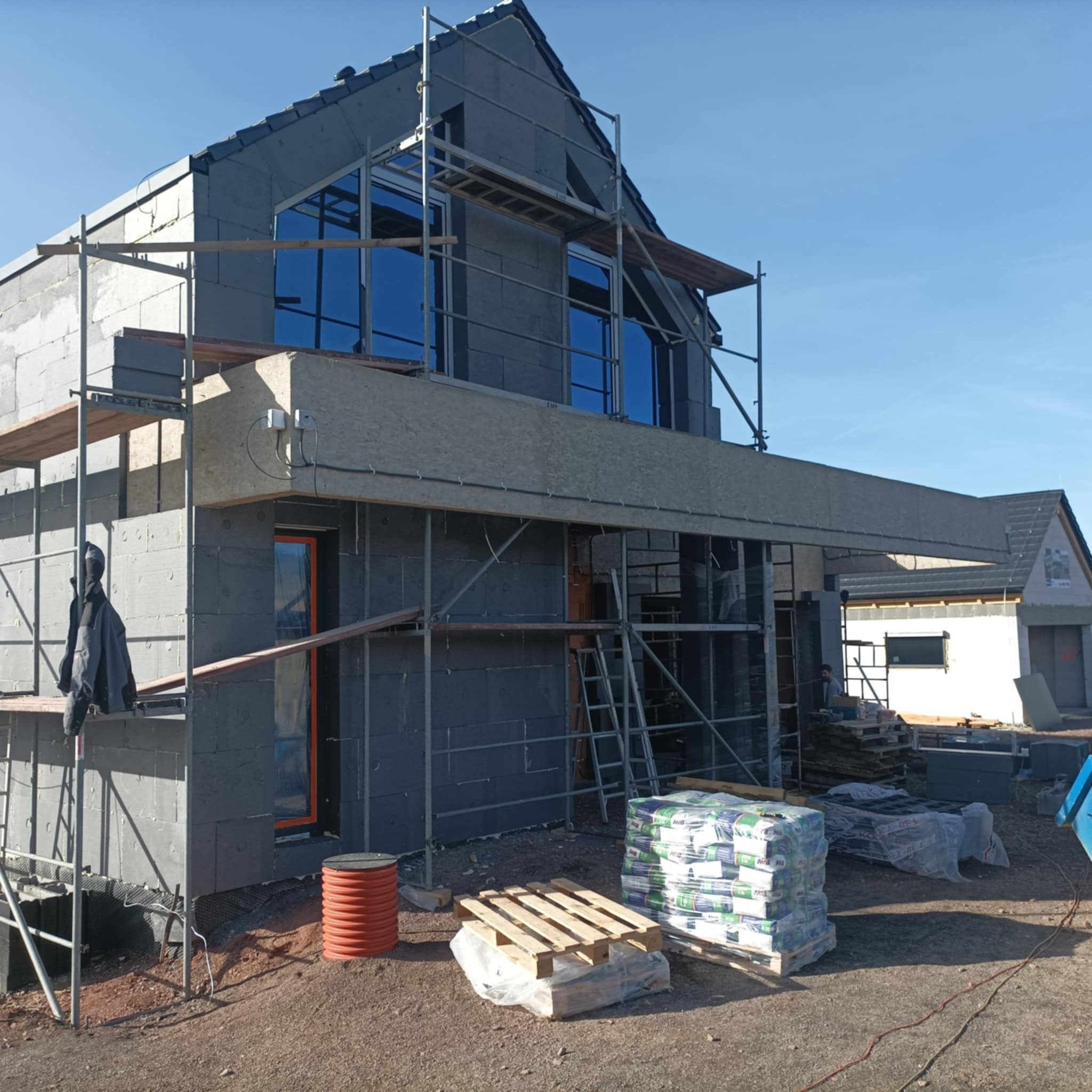 Dom w trakcie budowy, ocieplony styropianem, z zamontowanymi oknami, widoczne rusztowania, materiały budowlane i rury drenażowe na placu budowy.