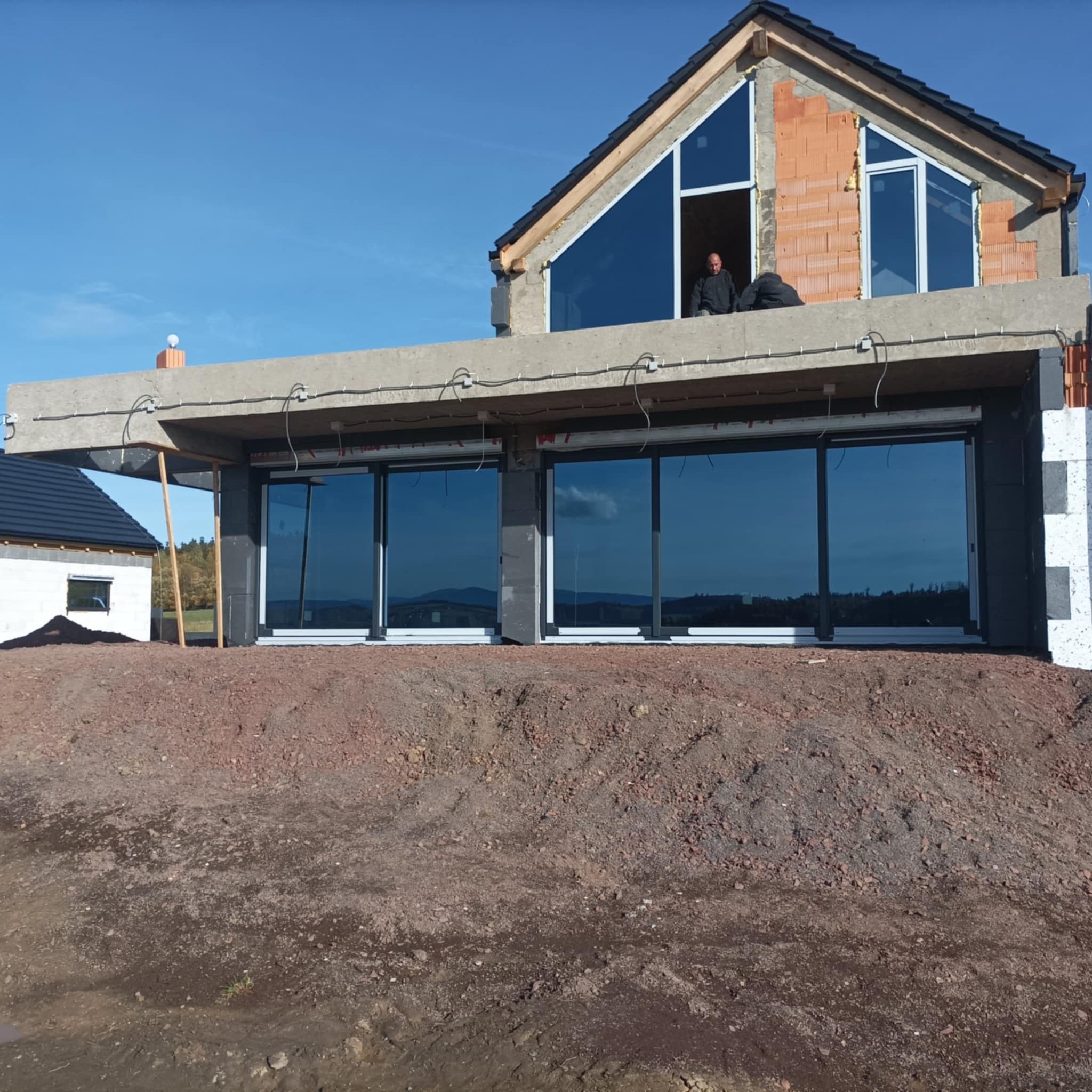 Budowa domu w stanie surowym otwartym: widoczne surowe ściany z cegły, duże okna z ciemnymi szybami, betonowy strop i dwaj pracownicy na piętrze.