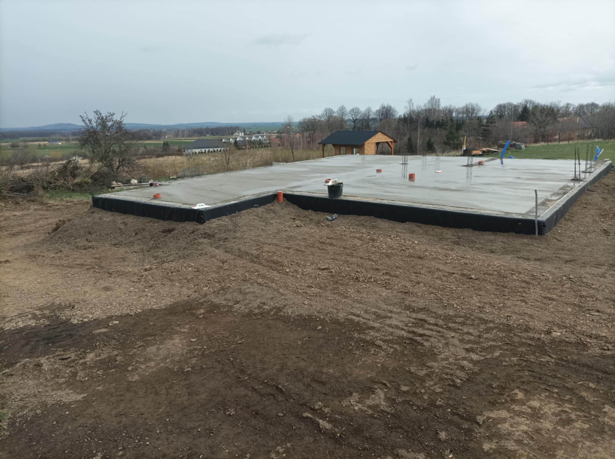 Wylany fundament płyty betonowej z widoczną hydroizolacją pionową, w tle krajobraz wiejski z nowo wybudowaną drewnianą altaną.