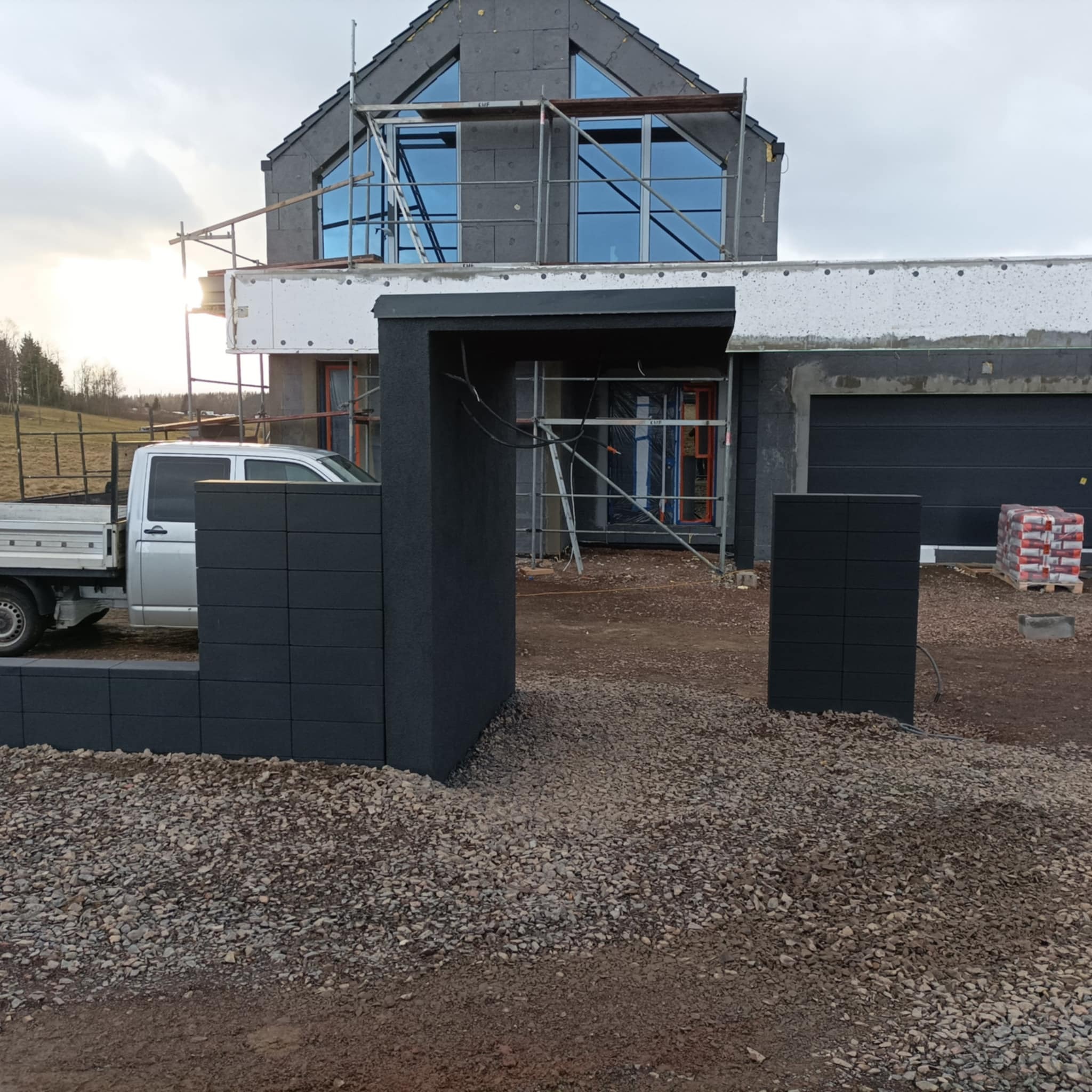 Nowoczesny dom w trakcie budowy z szarą elewacją, widoczne rusztowania, okna z niebieskimi szybami, częściowo wykonane ogrodzenie z szarych betonowych elementów, na podwórku leżą worki...