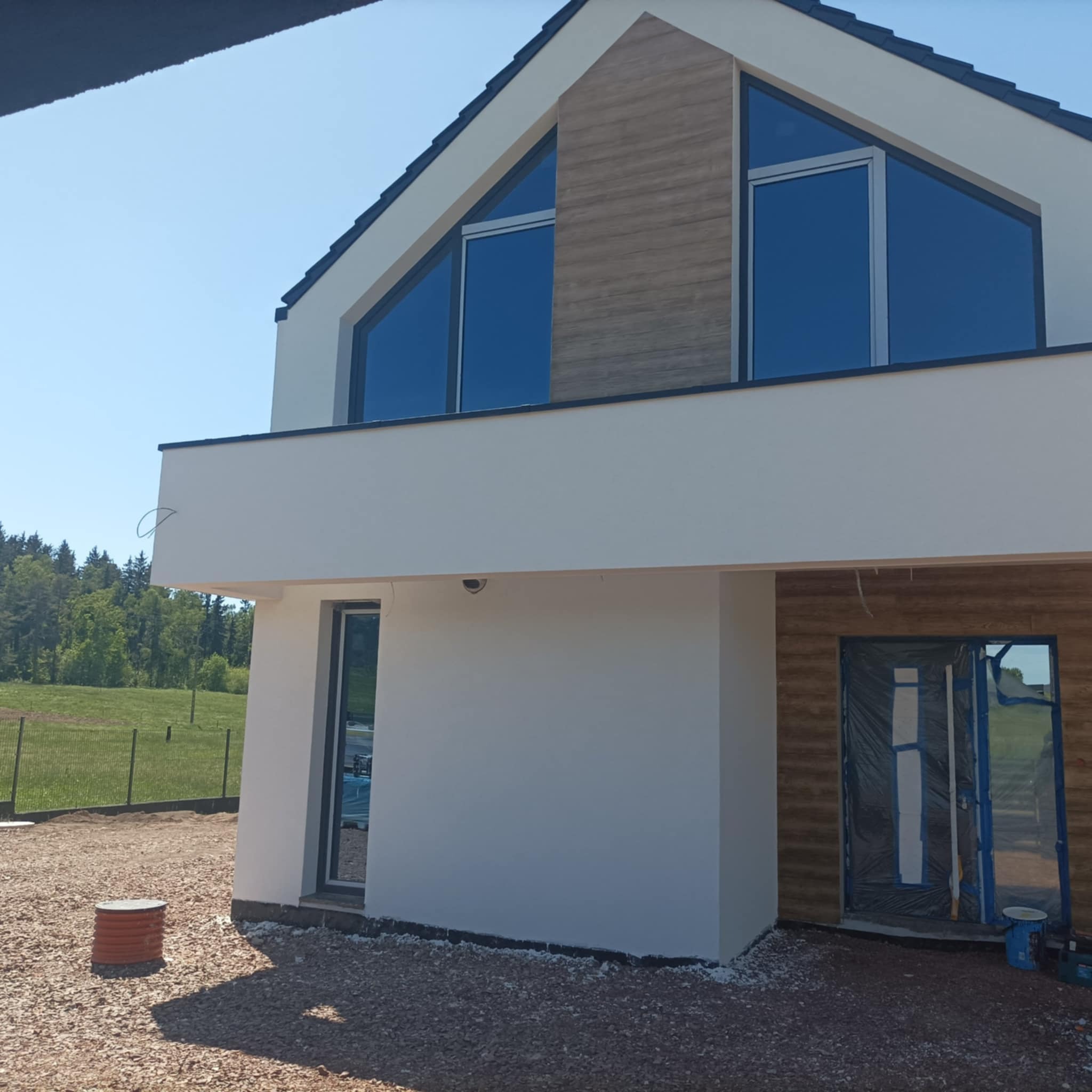 Nowoczesny dom w trakcie budowy, z elewacją w kolorze białym i drewnopodobnym, zabezpieczone drzwi wejściowe folią malarską, widoczny fragment drenażu i teren wokół domu wysypany żwirem.
