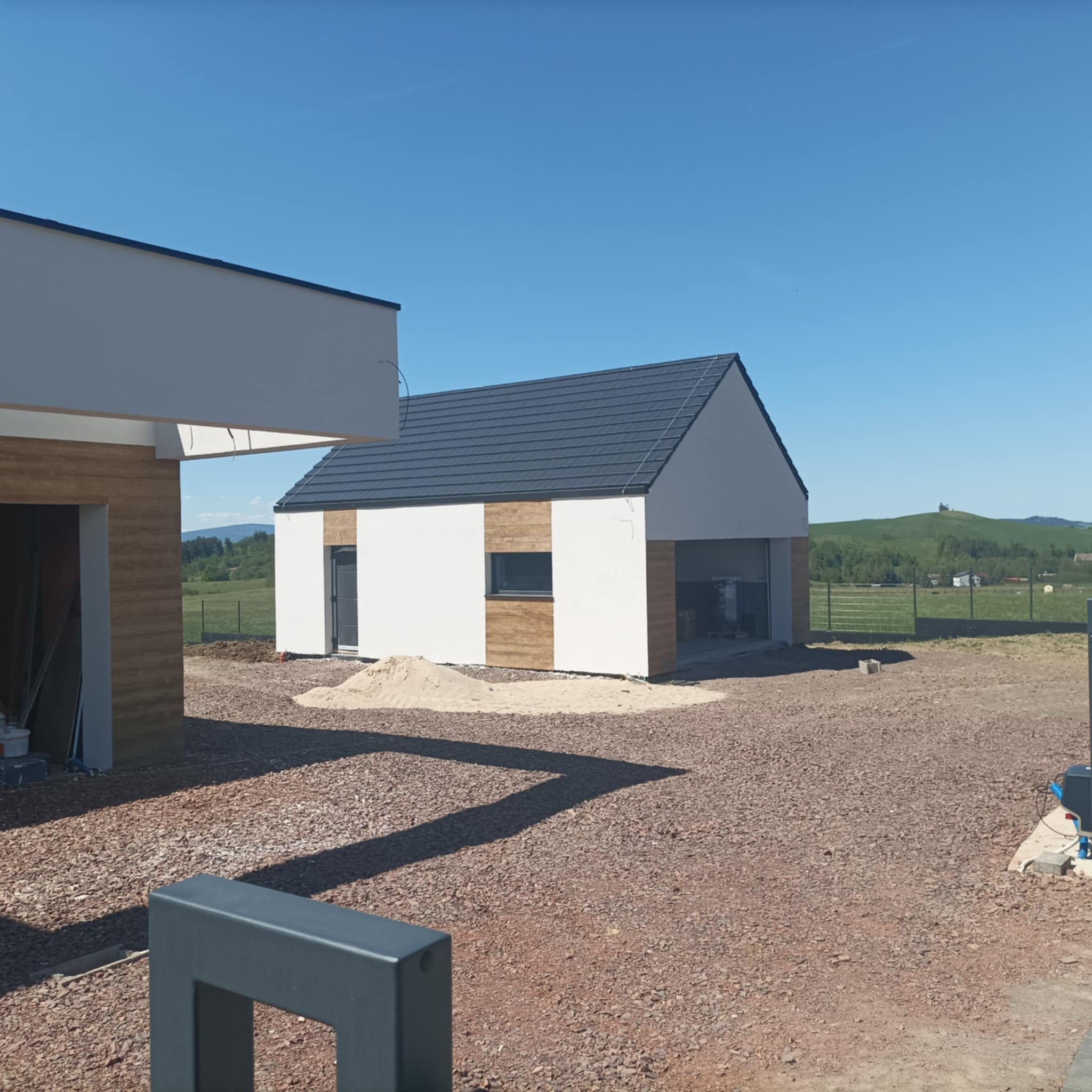 Nowoczesny dom z garażem w trakcie budowy, widoczny stos piasku, teren wokół domu wysypany żwirem, w tle pagórkowaty krajobraz.