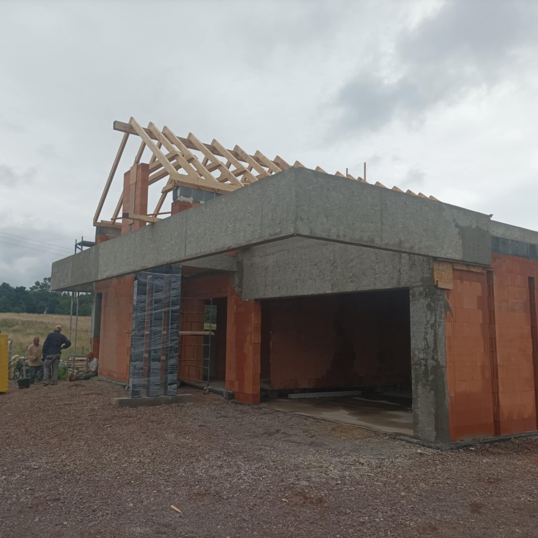 Budowa domu murowanego z czerwonej cegły, widoczne drewniane elementy konstrukcji dachu, betonowe nadproża i pracownicy w tle.