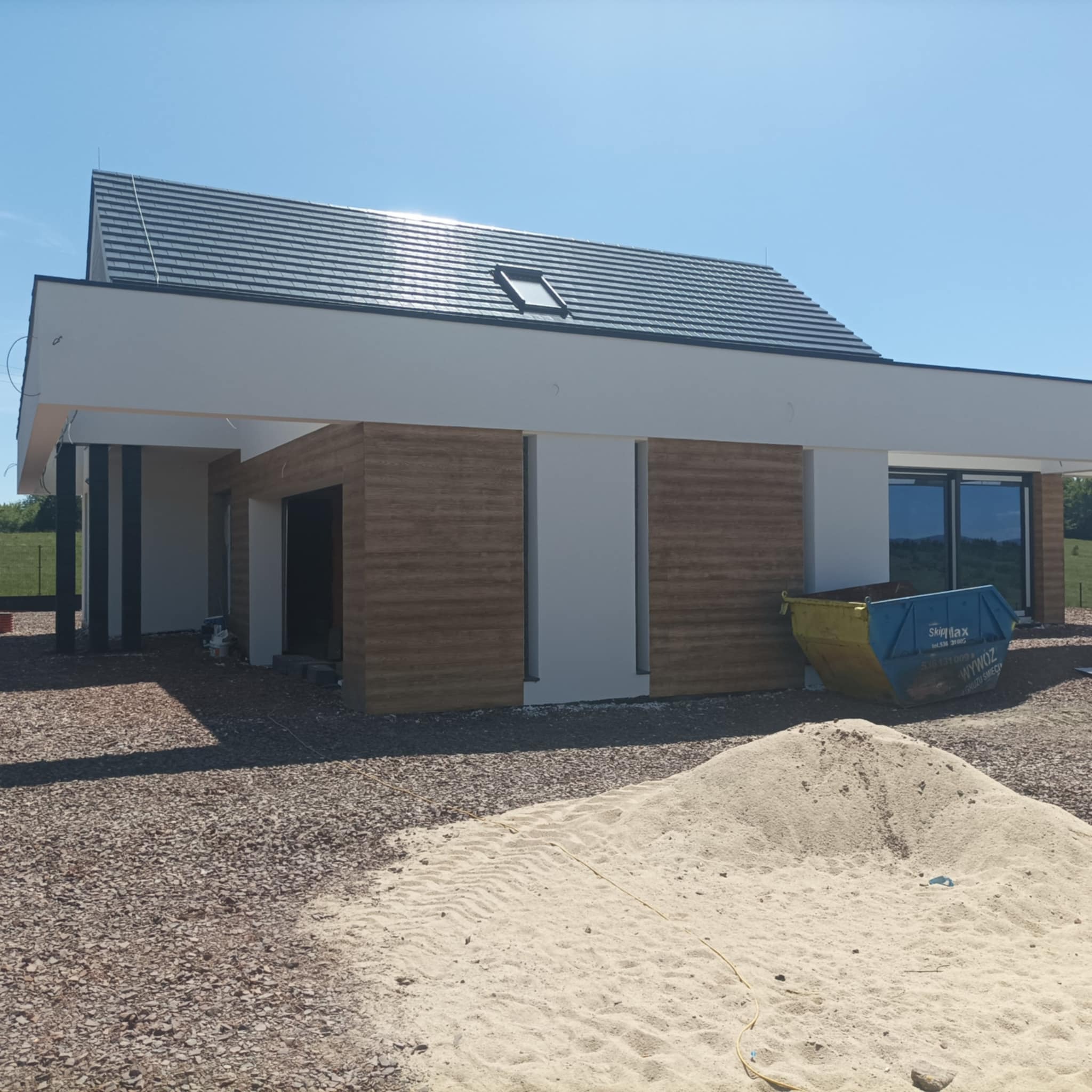 Nowoczesny dom jednorodzinny w trakcie budowy, z widoczną elewacją imitującą drewno, stertą piasku i kontenerem na gruz przed budynkiem, w słoneczny dzień.