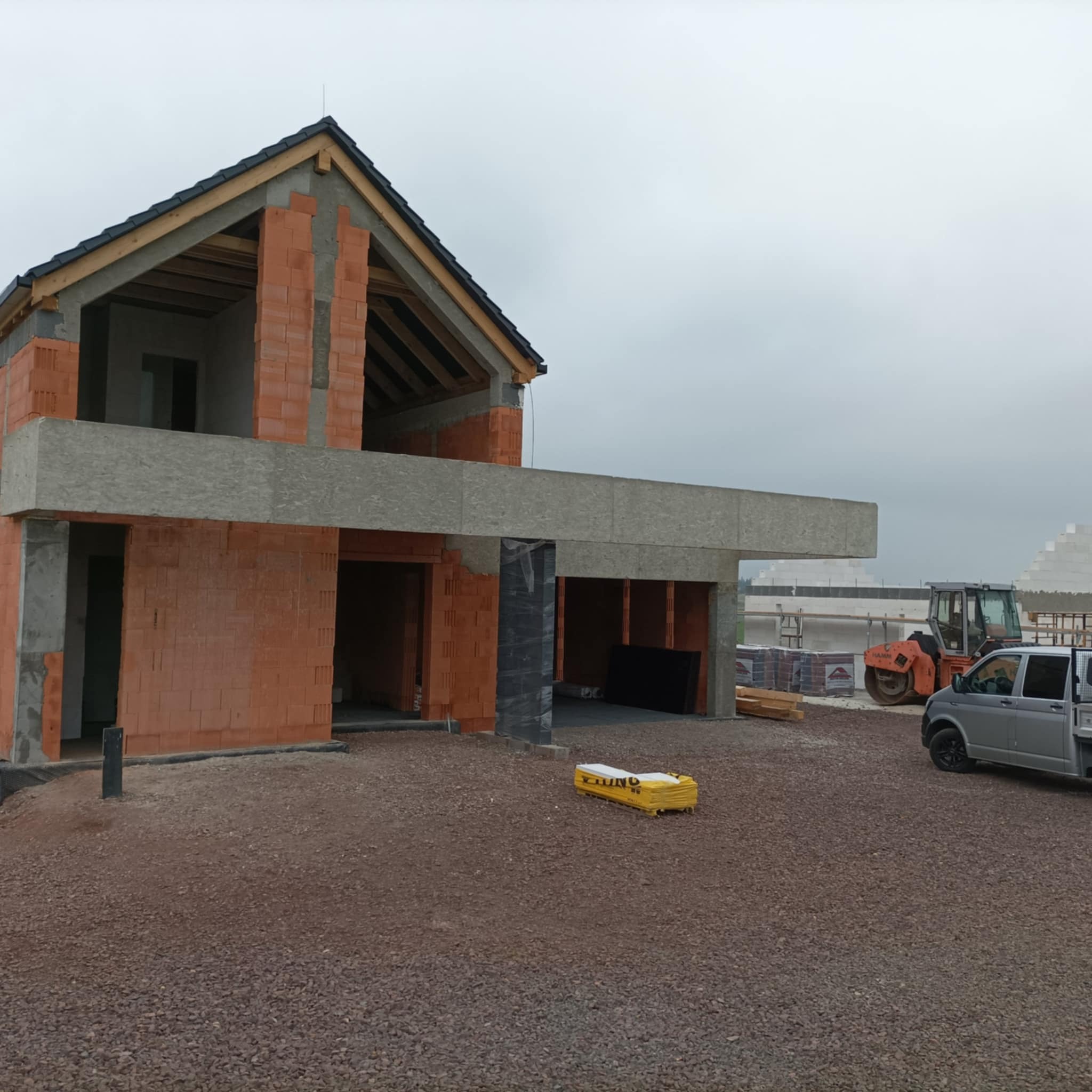 Surowy stan domu w trakcie budowy z czerwonej cegły, widoczny dach, brak okien, na posesji zaparkowana szara furgonetka i pomarańczowy walec drogowy, teren wysypany żwirem.