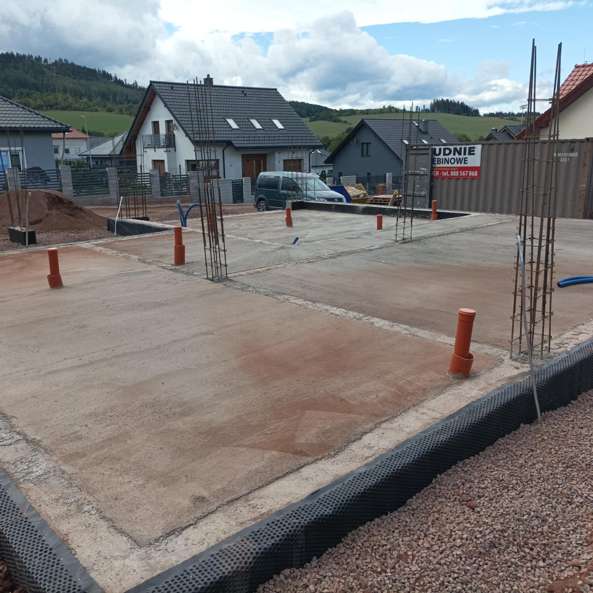 Zalany fundament z wystającymi prętami zbrojeniowymi i pomarańczowymi rurami, widoczne warstwy izolacyjne i podsypka żwirowa, w tle zabudowa jednorodzinna i zalesione wzgórza.