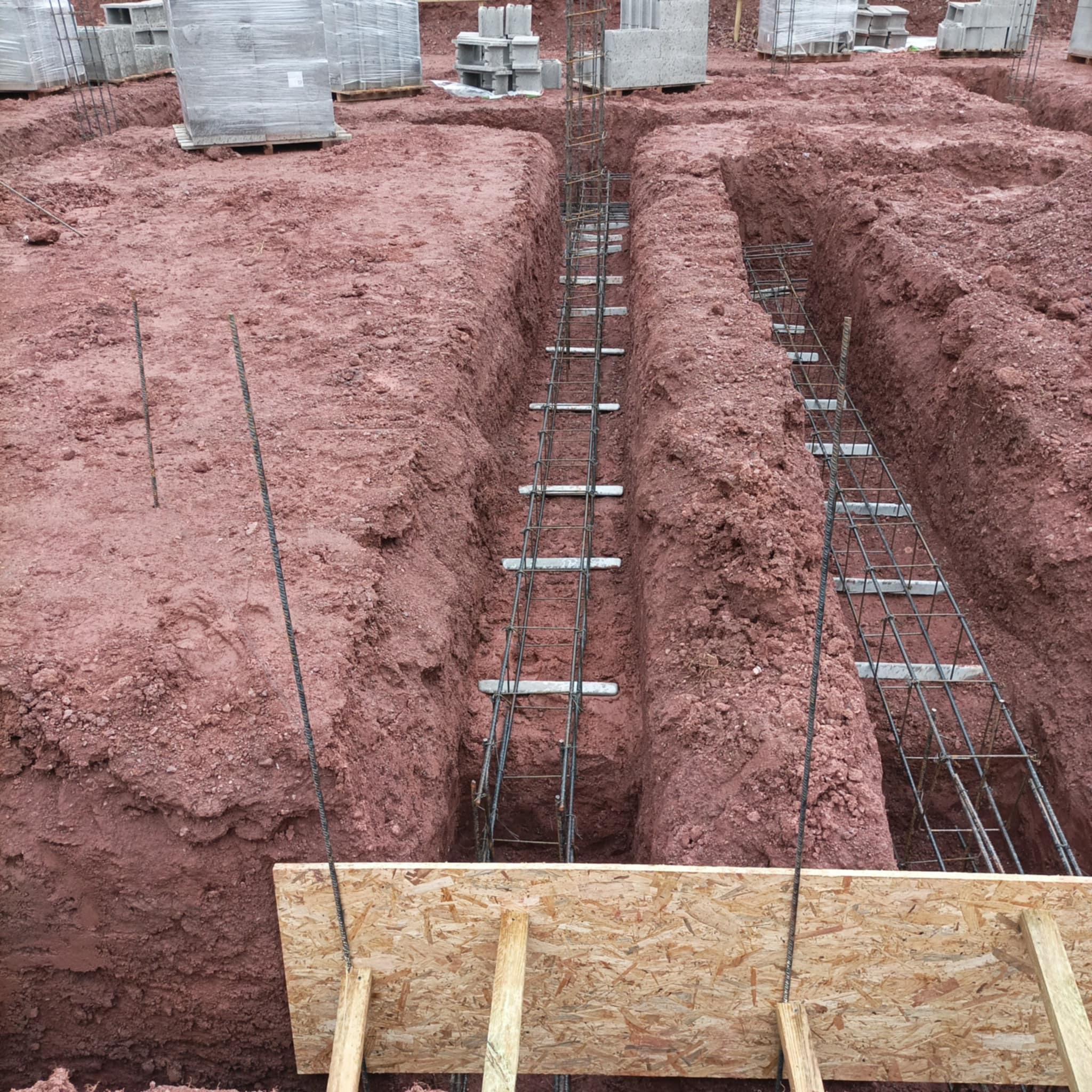 Przygotowanie zbrojenia w wykopach pod fundamenty, widoczne deskowanie i zapasy bloczków betonowych w tle na czerwonej ziemi.