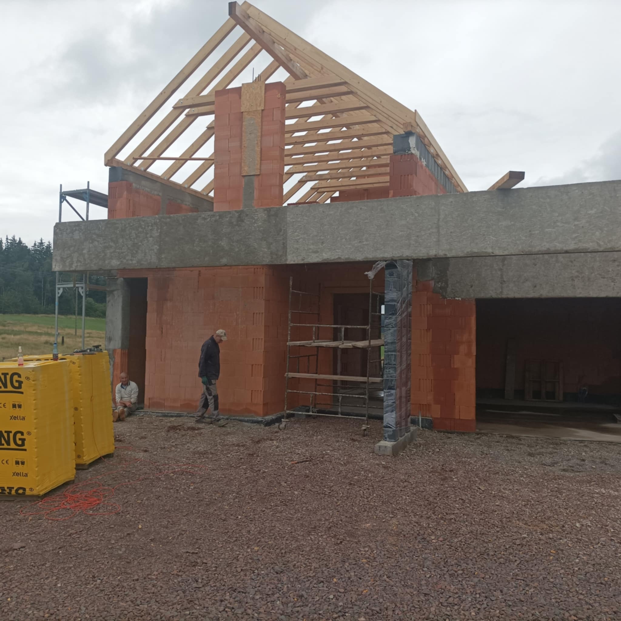 Budowa domu w trakcie wznoszenia ścian z czerwonej cegły, widoczne drewniane elementy konstrukcji dachu, rusztowania i materiały budowlane na placu budowy, dwóch robotników przy pracy.