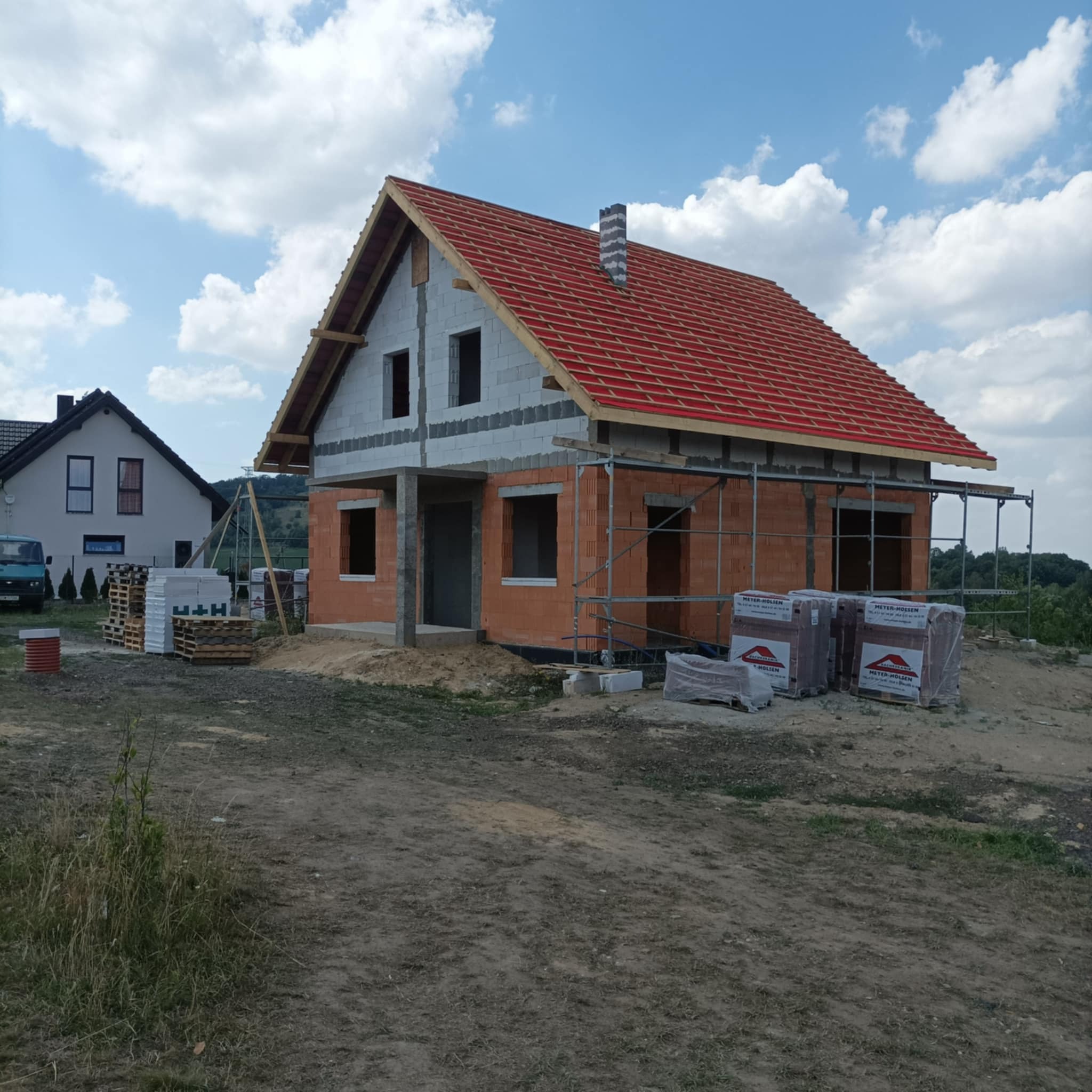 Budowa domu w trakcie realizacji: ściany z czerwonej cegły częściowo wymurowane, dach z czerwoną dachówką w trakcie układania, widoczne rusztowania i palety z materiałami budowlanymi, w tle inny...