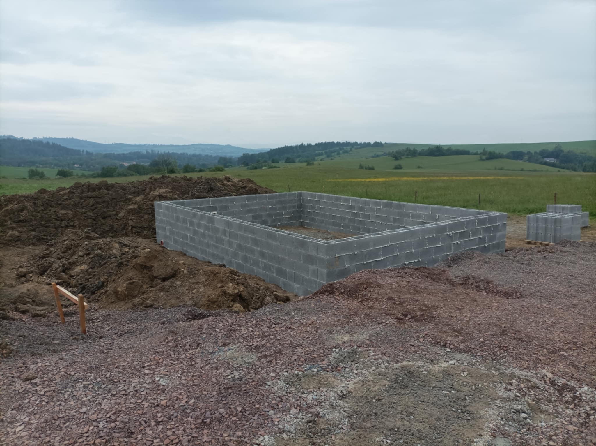 Wzniesiony fundament z bloczków betonowych na terenie budowy, widoczne pryzmy ziemi i oddalony krajobraz pagórkowaty pod pochmurnym niebem.