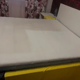 Biały materac na łóżku z żółtą narzutą, widoczne ślady po czyszczeniu, obok szafka nocna z butelką wody.