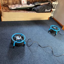 Pranie Tąpiecerki Clean_Service - Dwa wentylatory osuszające z niebieskimi elementami na ciemnoniebieskim dywanie po czyszczeniu, widok z góry, kabel zasilający na podłodze.