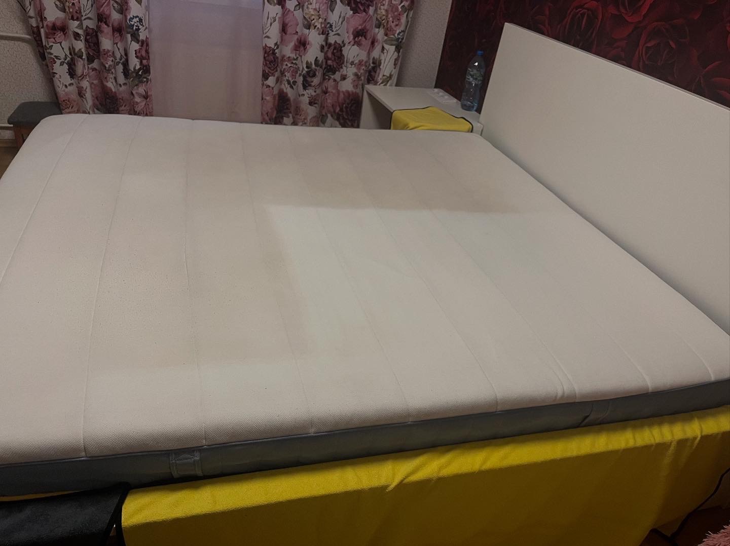 Biały materac na łóżku z żółtą narzutą, widoczne ślady po czyszczeniu, obok szafka nocna z butelką wody.