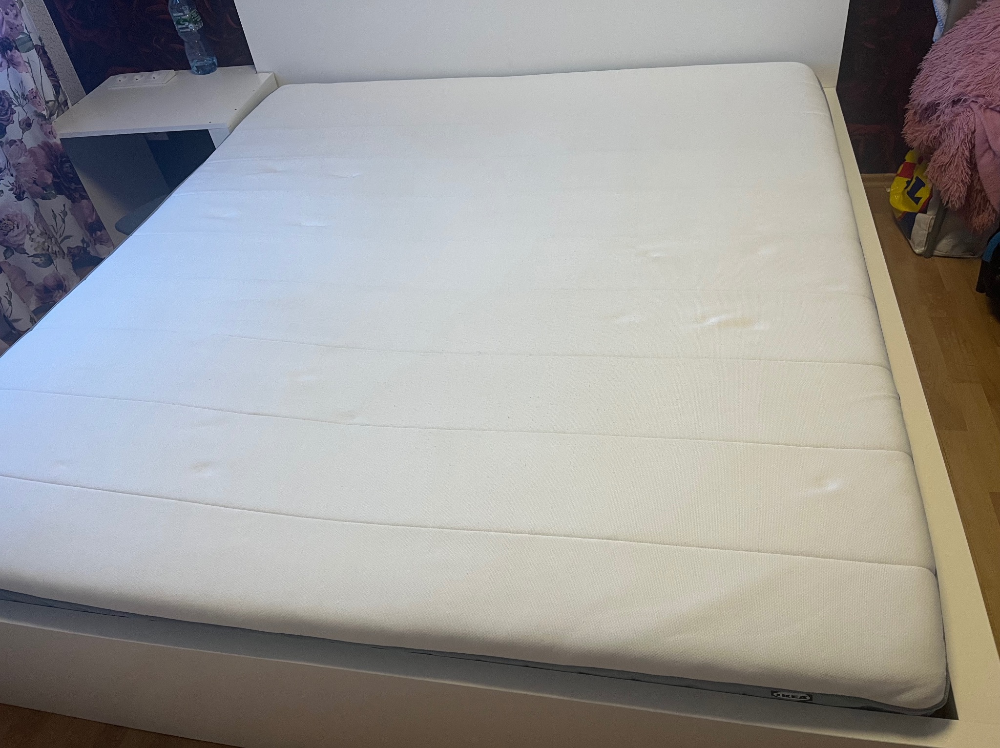 Biały materac z widocznymi plamami na powierzchni, leżący na białej ramie łóżka, z szafką nocną i butelką wody w tle.