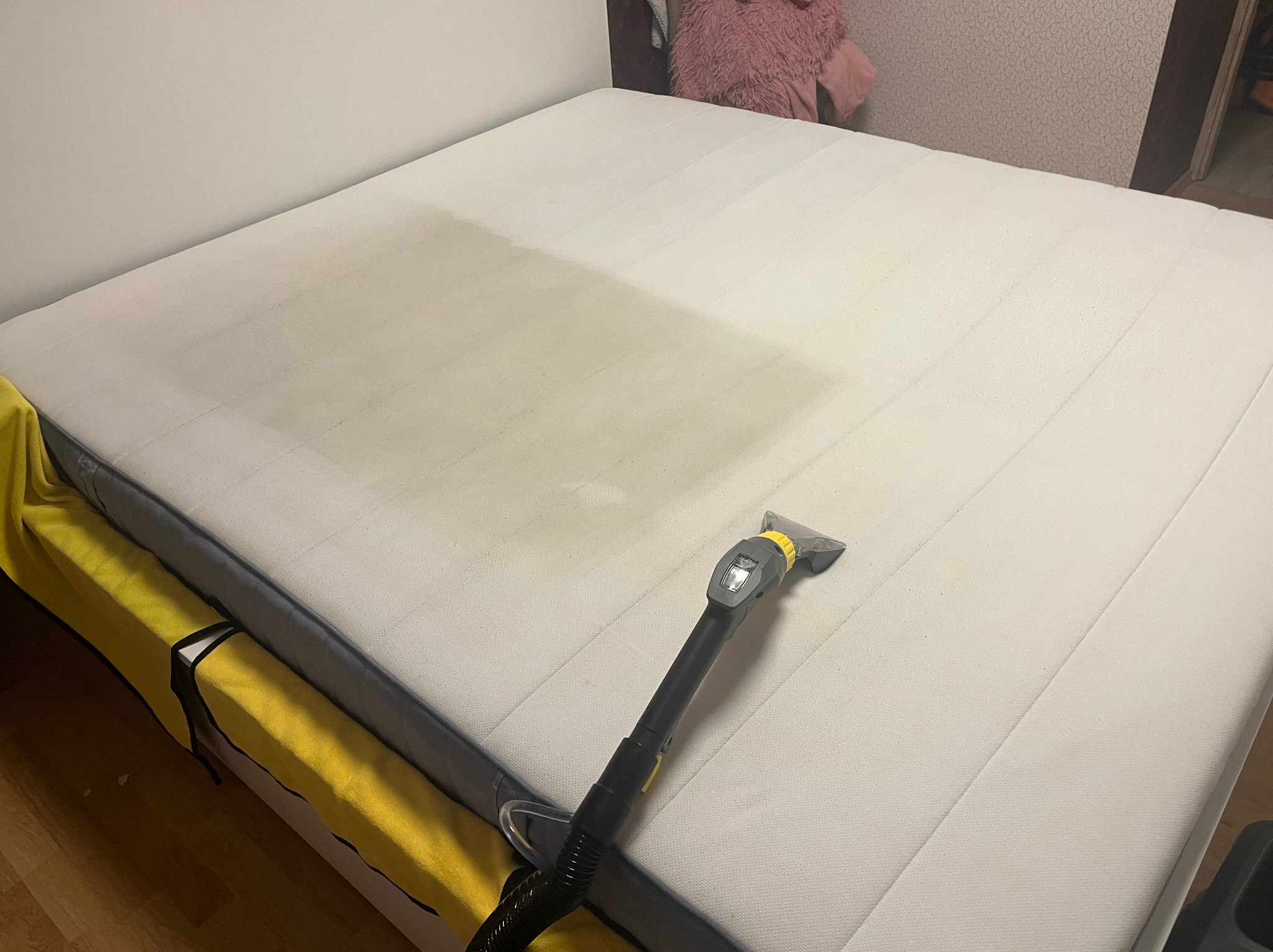 Czyszczenie materaca z widocznym zabrudzeniem i używanym odkurzaczem piorącym, zabezpieczone żółtą tkaniną boki łóżka.