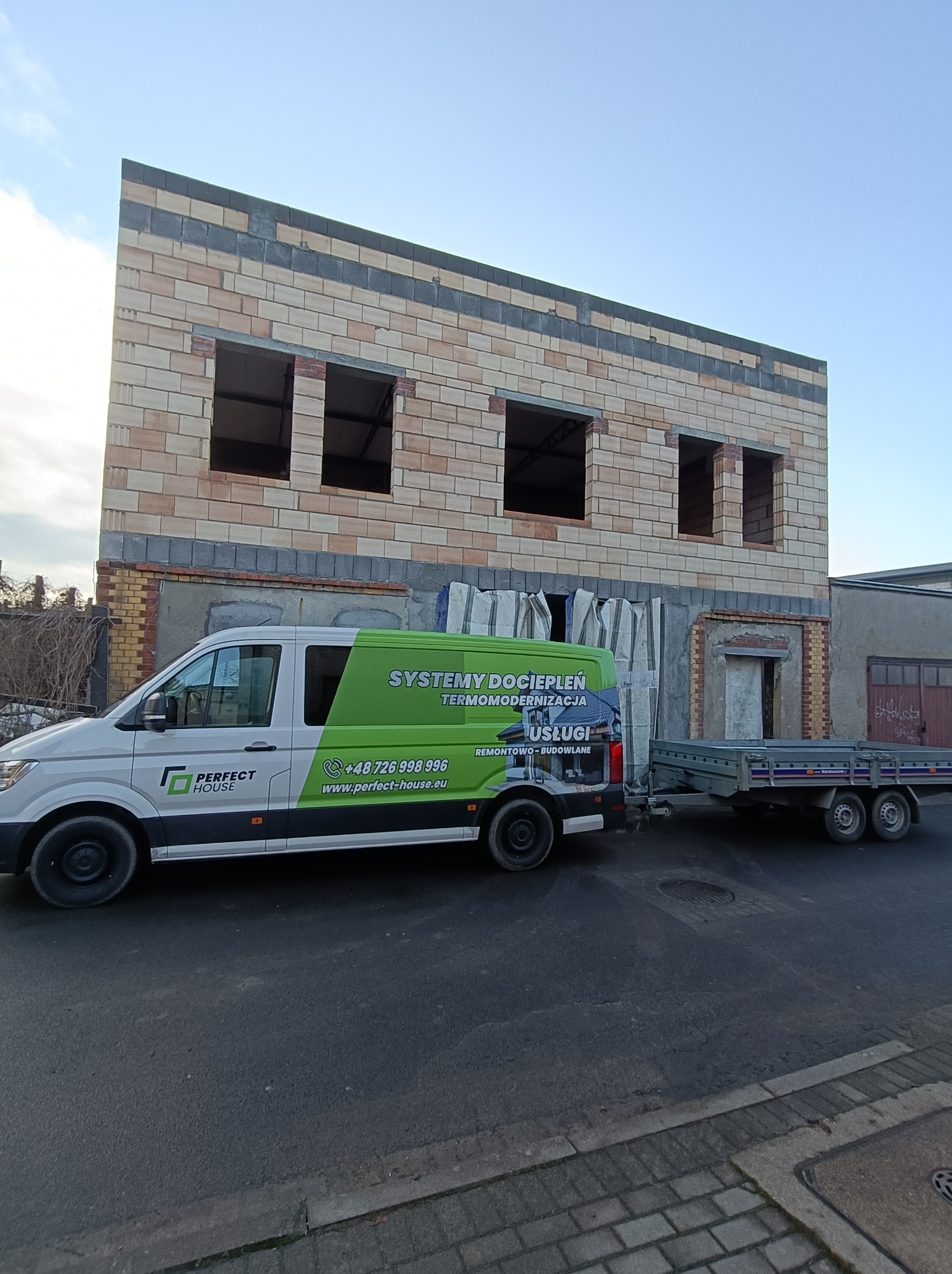 Dwukondygnacyjny budynek w trakcie murowania ścian z pustaków ceramicznych, widoczny z boku, z zaparkowanym firmowym vanem z przyczepką. Na budynku widoczne otwory okienne.