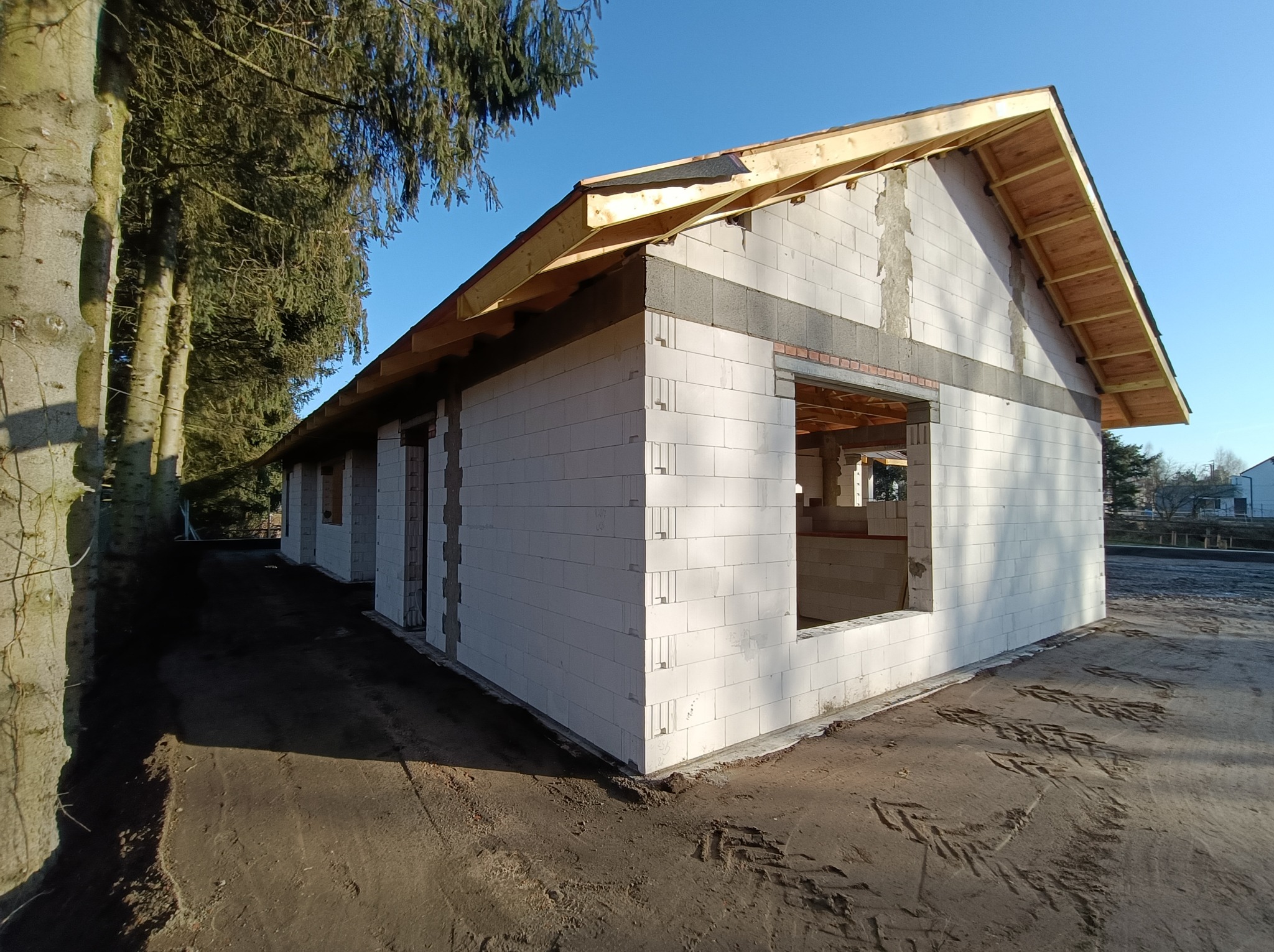 Surowy stan domu z białych bloczków, widoczne wnęki okienne i drewniana konstrukcja dachu, otoczenie z ziemi i drzew w tle.