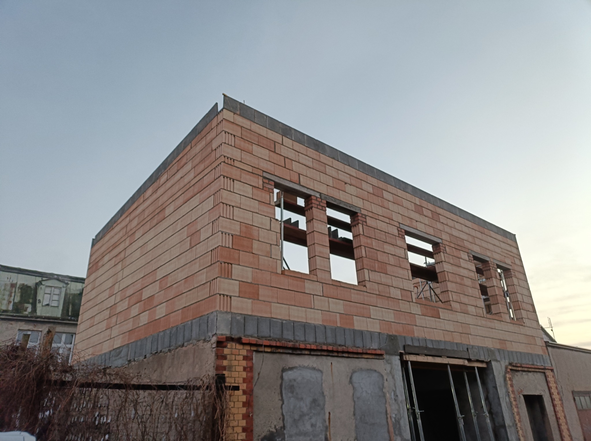 Niedokończony budynek z cegły ceramicznej, widoczne otwory okienne z drewnianymi belkami, dolna część budynku z elementami ceglanymi i betonowymi, w tle inny budynek.