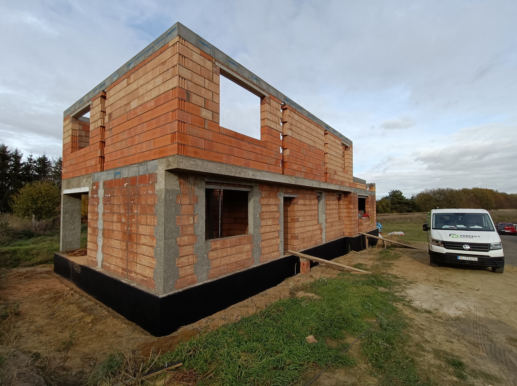 Wznoszenie ścian domu z pustaków ceramicznych, widoczna czarna izolacja fundamentów i firmowy bus 'Perfect House' w tle.