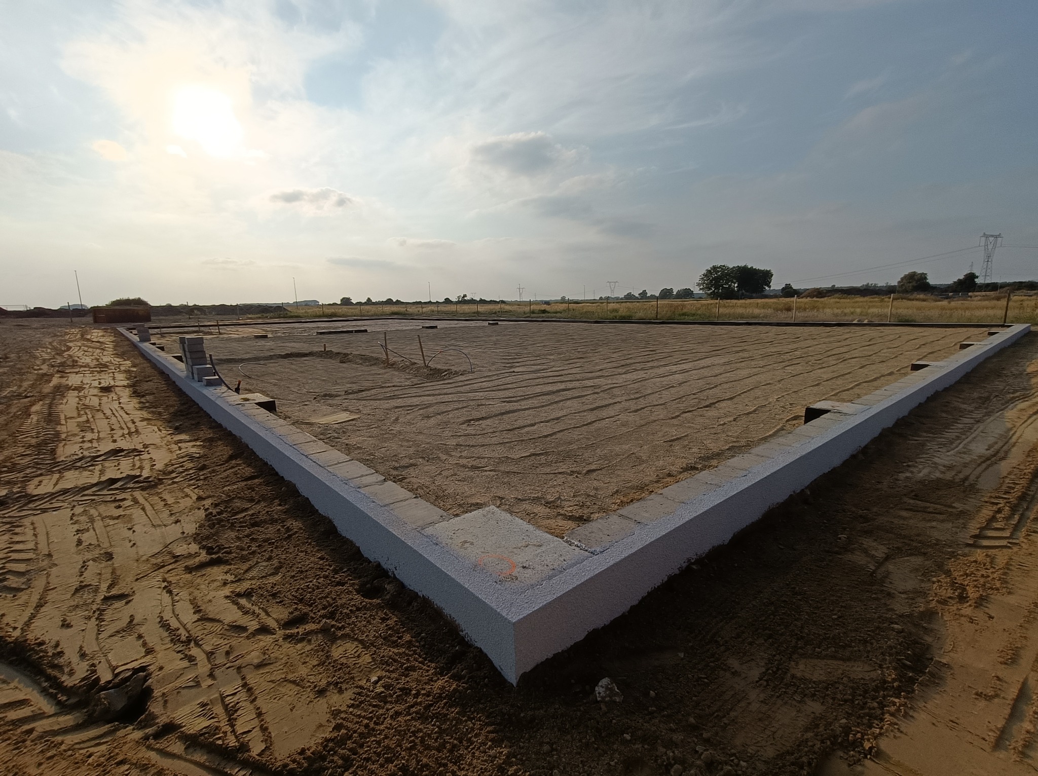 Fundamenty domu jednorodzinnego w trakcie budowy, z widocznymi bloczkami fundamentowymi tworzącymi obrys budynku na przygotowanym podłożu piaskowym, w tle widoczne słupy wysokiego napięcia.