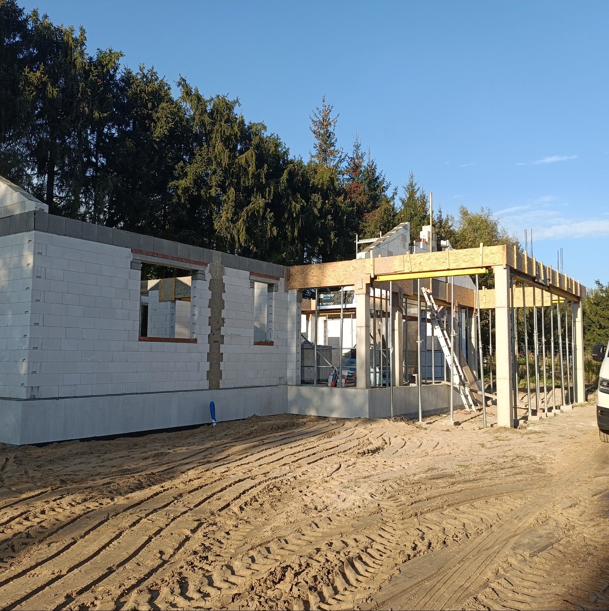 Budowa domu w Stęszewie w trakcie wznoszenia ścian z bloczków i konstrukcji żelbetowej, widoczne ślady opon na piaszczystym gruncie.