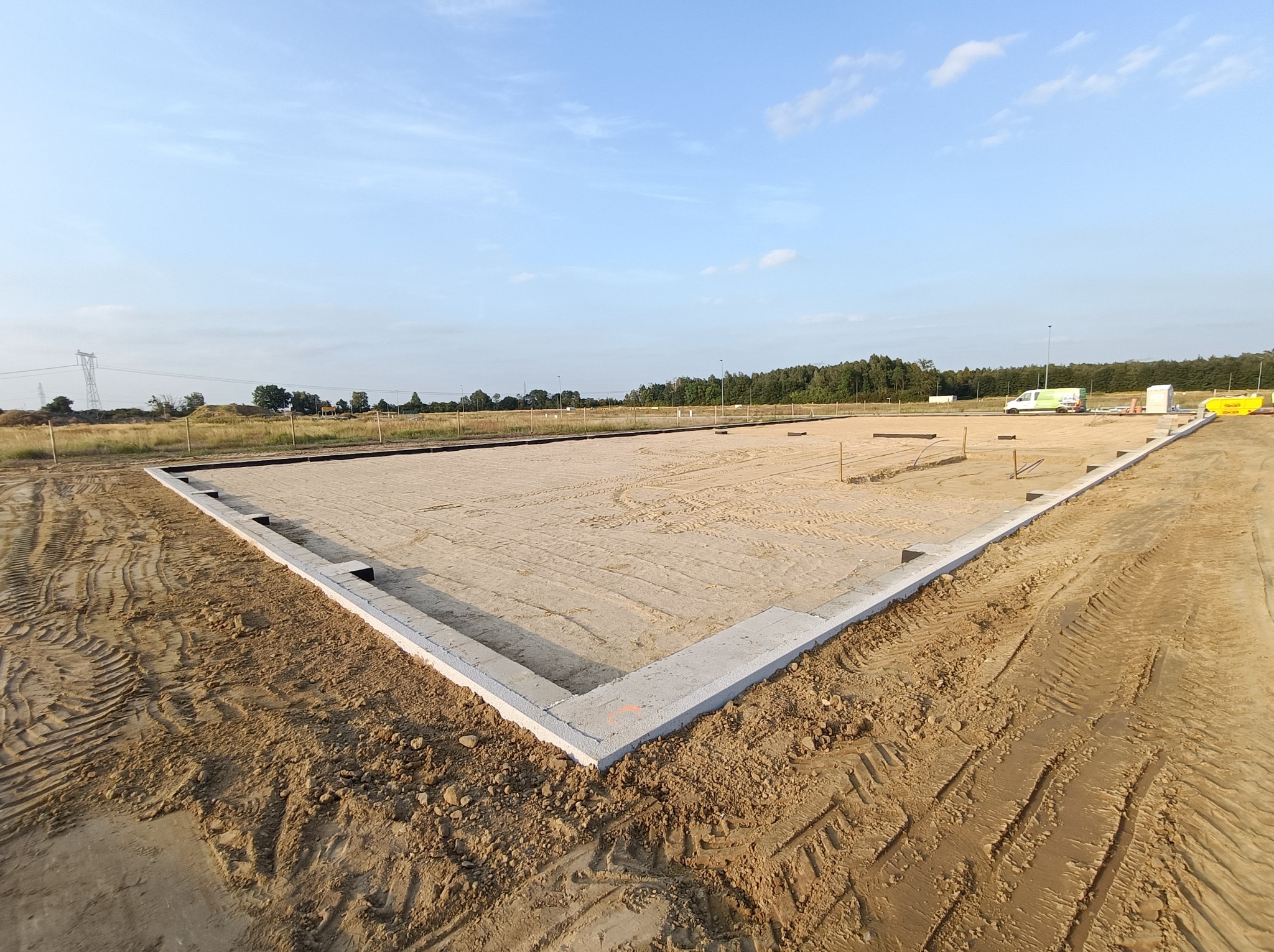 Przygotowany teren pod budowę domu z wylanymi betonowymi fundamentami, widoczne ślady opon na piasku, w tle drzewa i błękitne niebo.