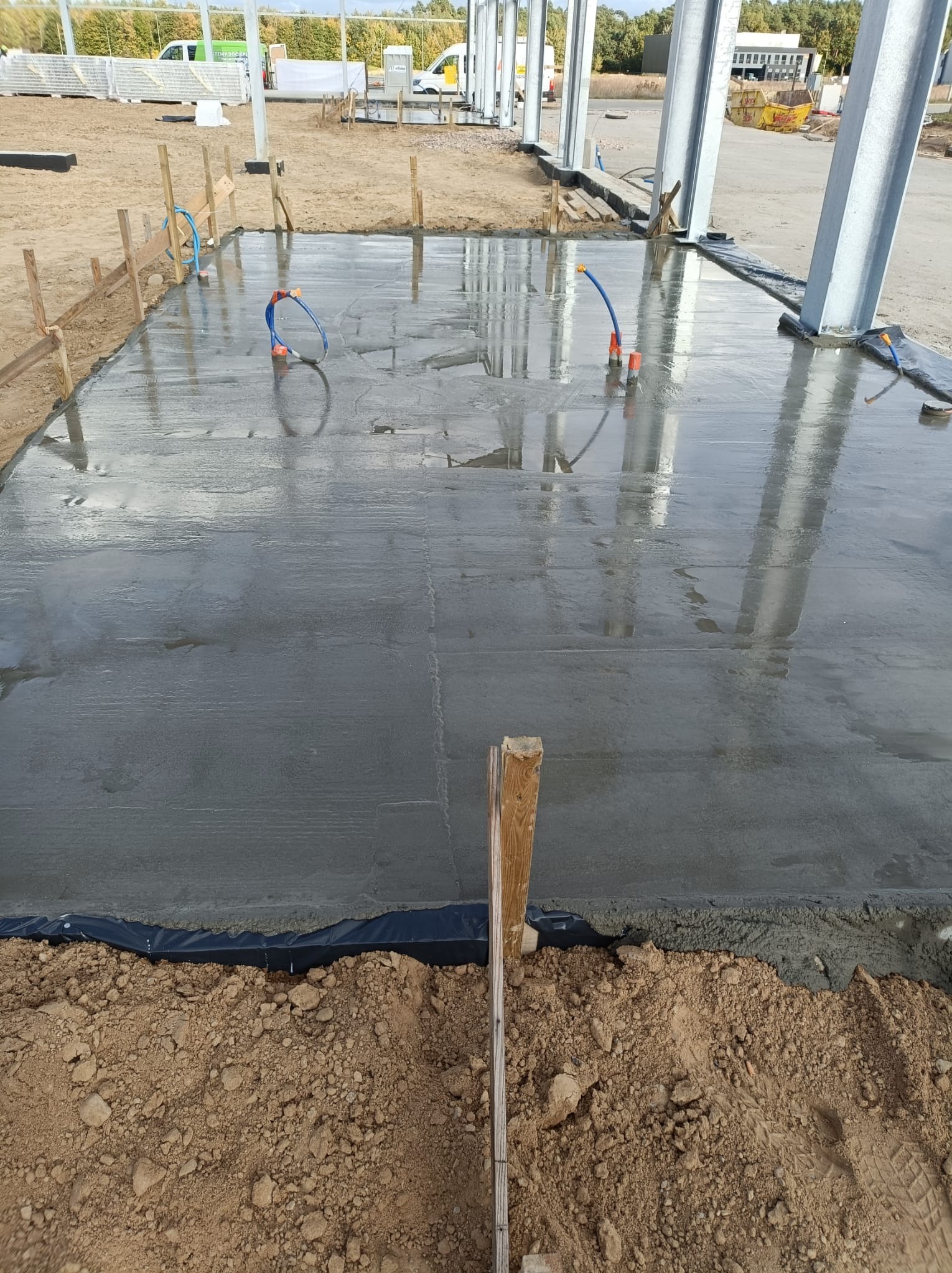 Świeżo wylana, mokra płyta betonowa z widocznymi instalacjami i drewnianym szalunkiem, przygotowana pod budowę hali stalowej, z widocznym gruntem wokół