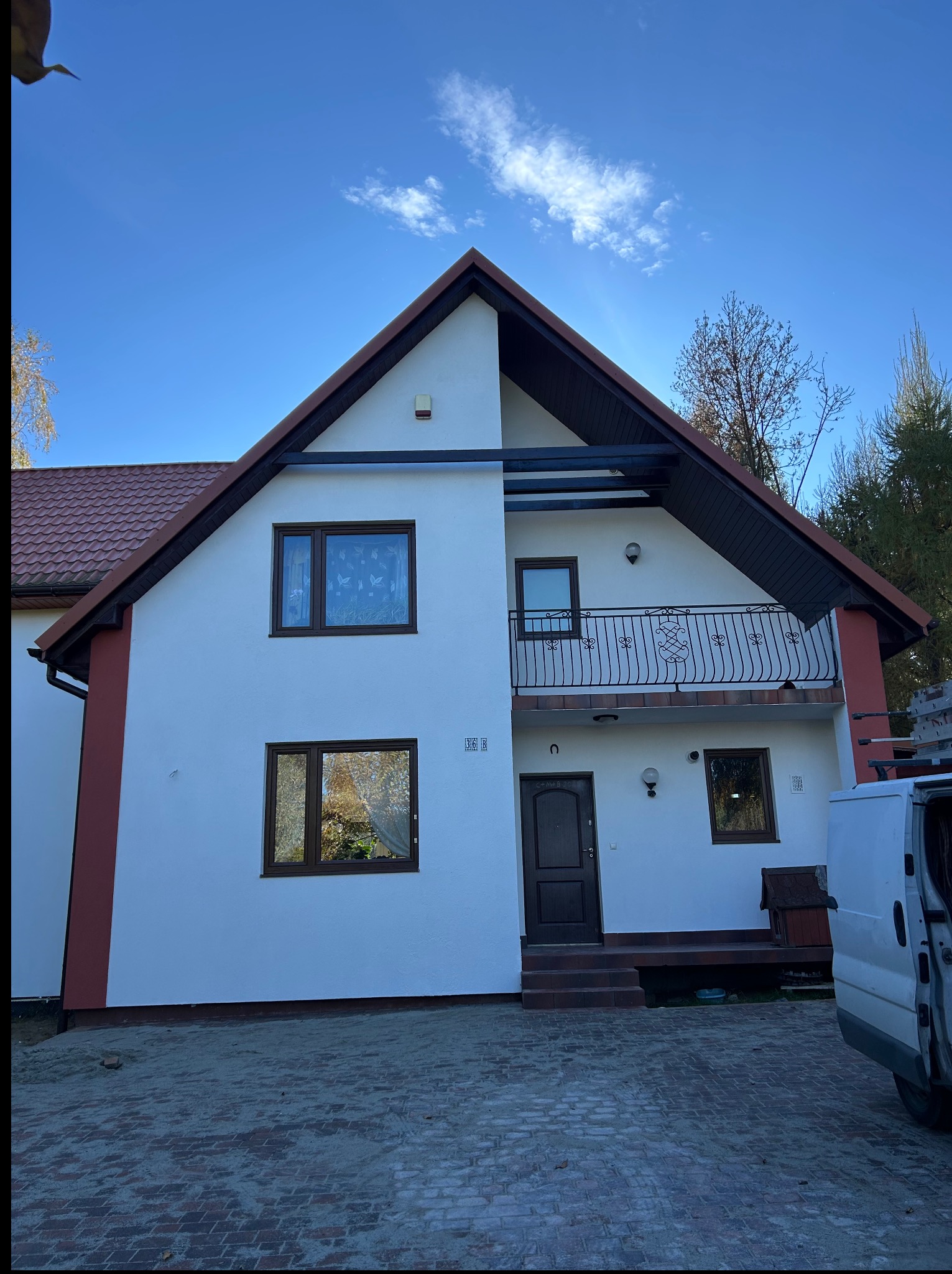 Biała elewacja domu jednorodzinnego z brązowymi oknami i dachem, po malowaniu. Widoczny fragment samochodu dostawczego i kostka brukowa na podjeździe.