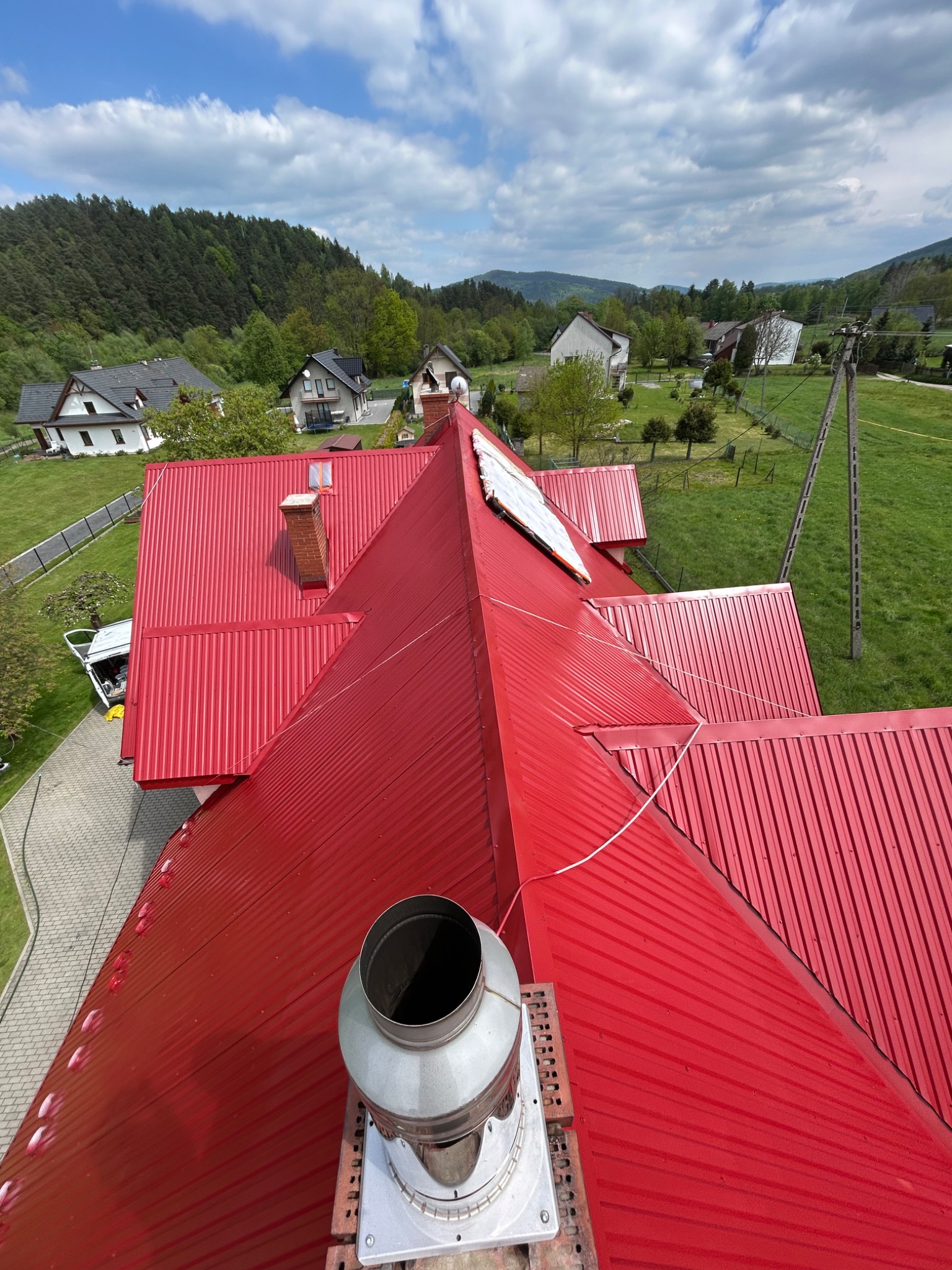 Czerwony, blaszany dach domu z kominem i wentylacją, widziany z góry na tle zielonych wzgórz i błękitnego nieba. Widoczne detale blacharskie i elementy konstrukcyjne dachu.