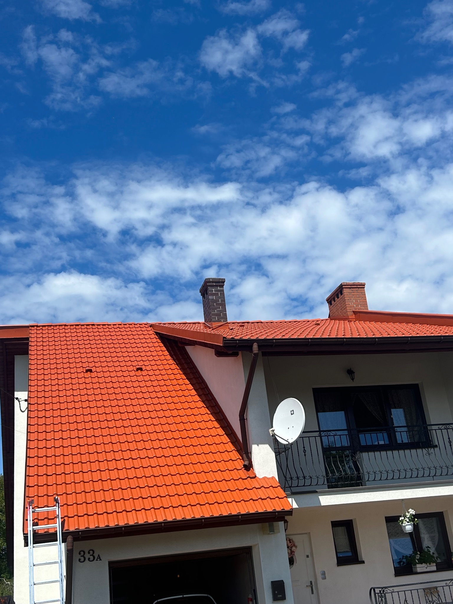 Dom z czerwoną dachówką i drabiną opartą o rynnę. Widoczne błękitne niebo z chmurami. Ujęcie z dołu podkreśla wysokość budynku i detale dachu.