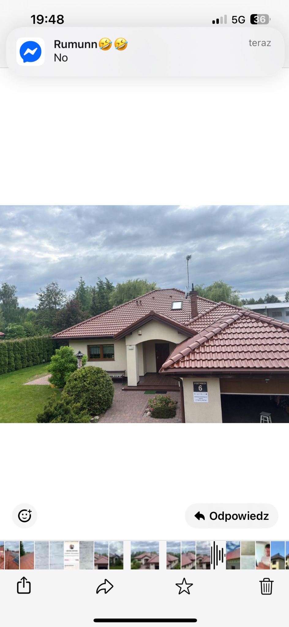 Dom jednorodzinny z czerwonym dachem i beżową elewacją, widok z góry. Na ścianie numer domu. Otoczenie: trawnik, drzewa, krzewy. Pochmurne niebo.