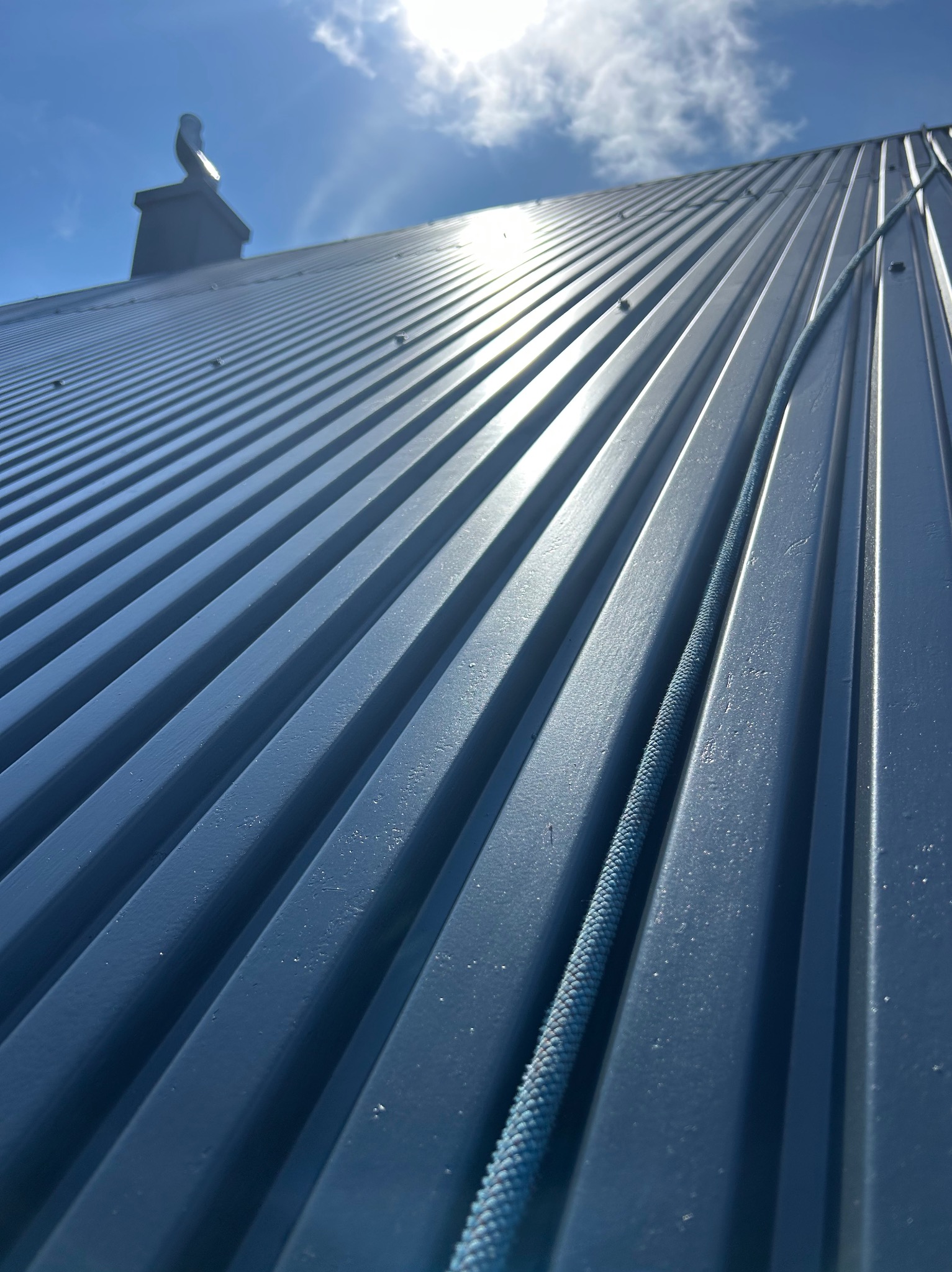 Błękitny, metalowy dach z kominem w słoneczny dzień, widziany z dołu pod kątem. Na dachu leży niebieska lina. Widoczne chmury na niebie.