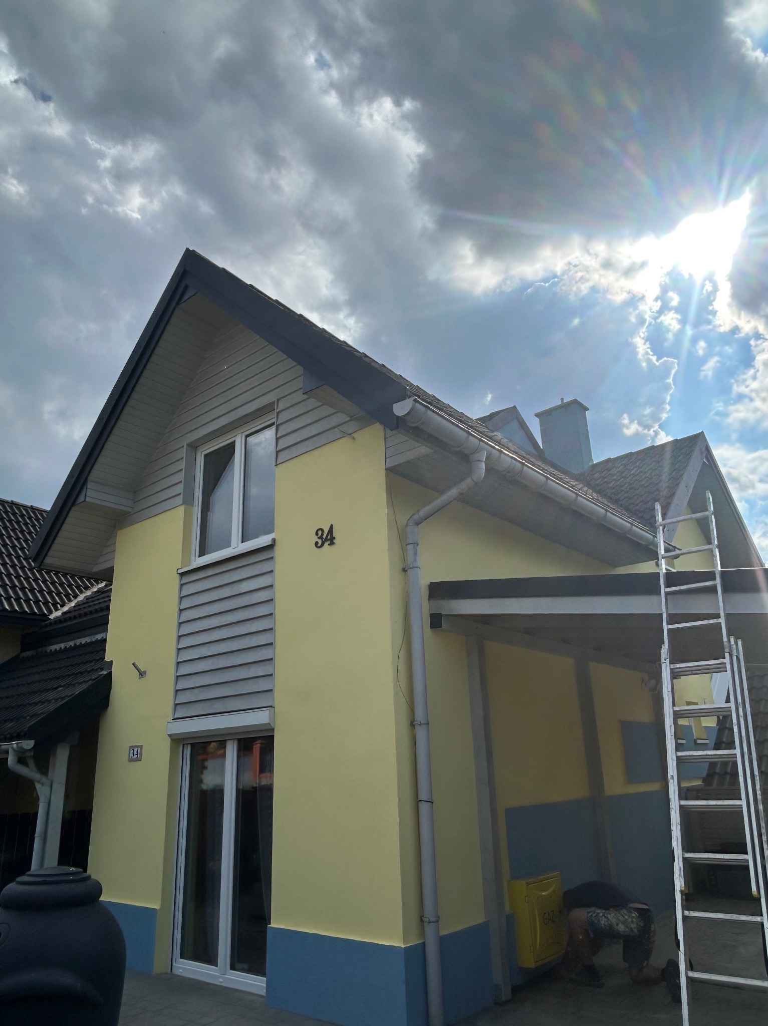 Żółta elewacja domu z szarą stolarką i numerem 34, widoczna drabina i fragment pracującego człowieka. Słoneczny dzień, częściowo zachmurzone niebo.