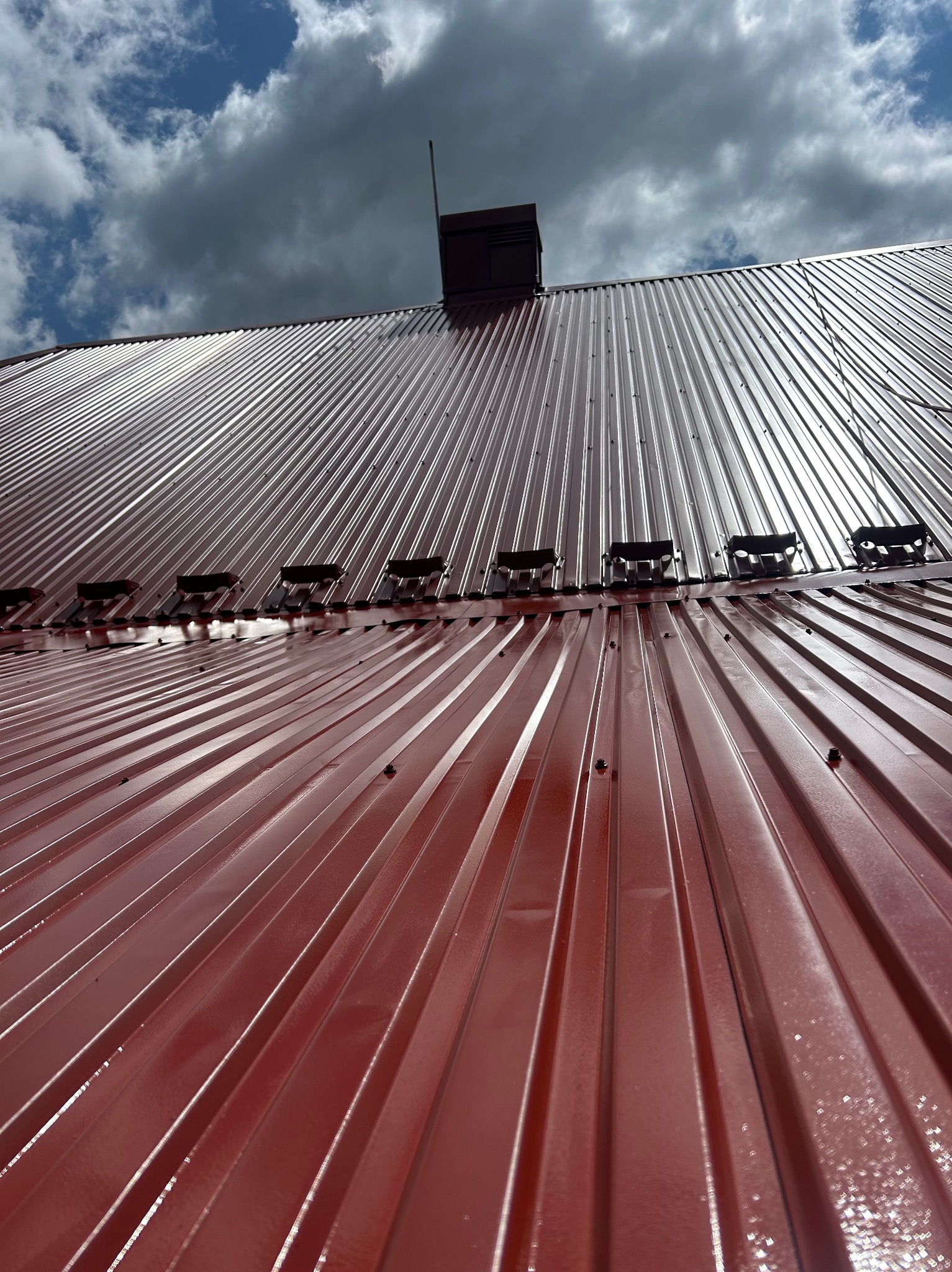Błyszczący, czerwony dach z blachy trapezowej, widok z dołu, fragment komina i anteny na tle pochmurnego nieba. Widoczne mocowania.