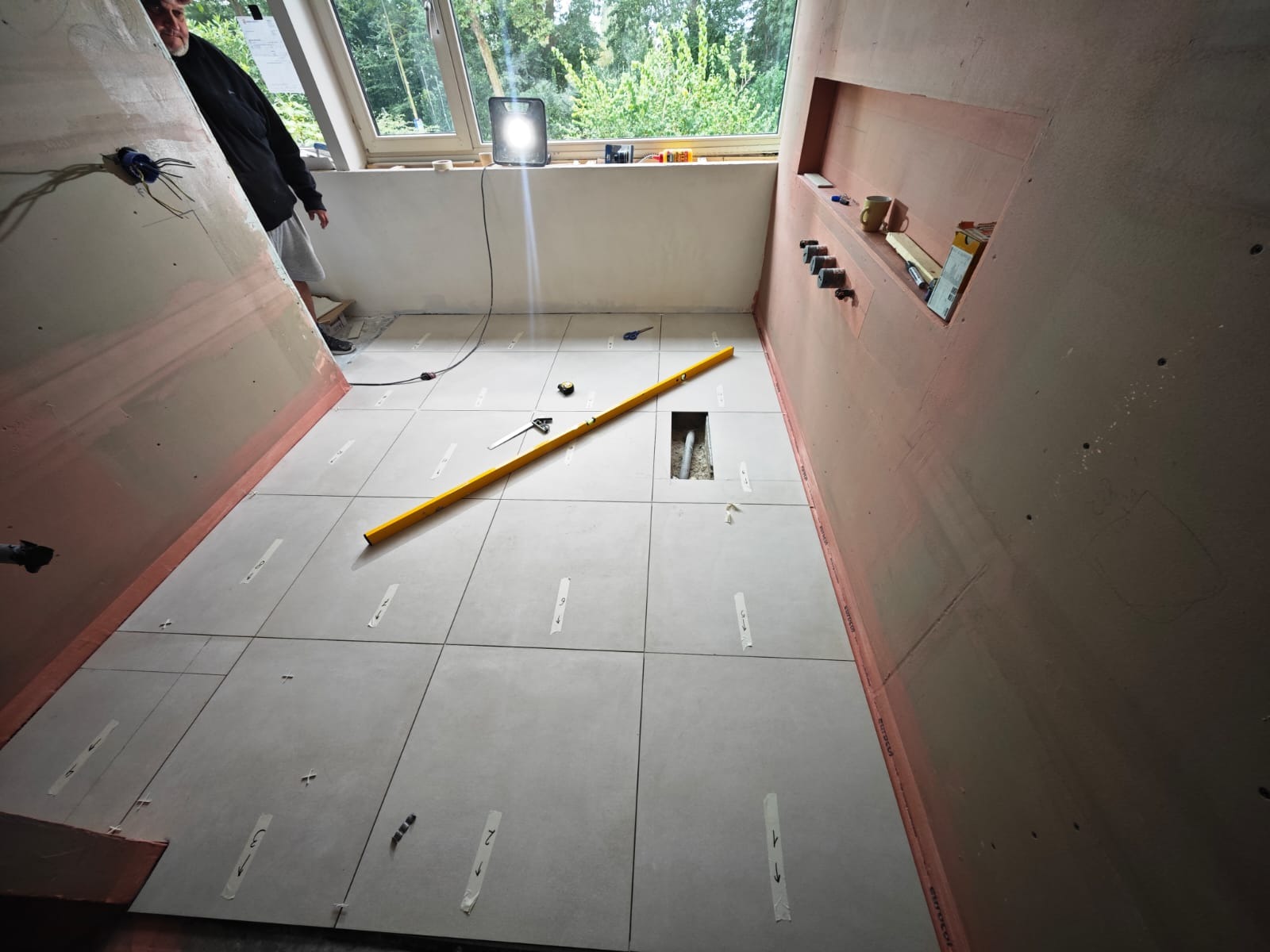 Układanie szarych płytek na podłodze w trakcie prac, widoczna poziomica, taśma malarska z numeracją, oraz przygotowane ściany i instalacja hydrauliczna w niszach.