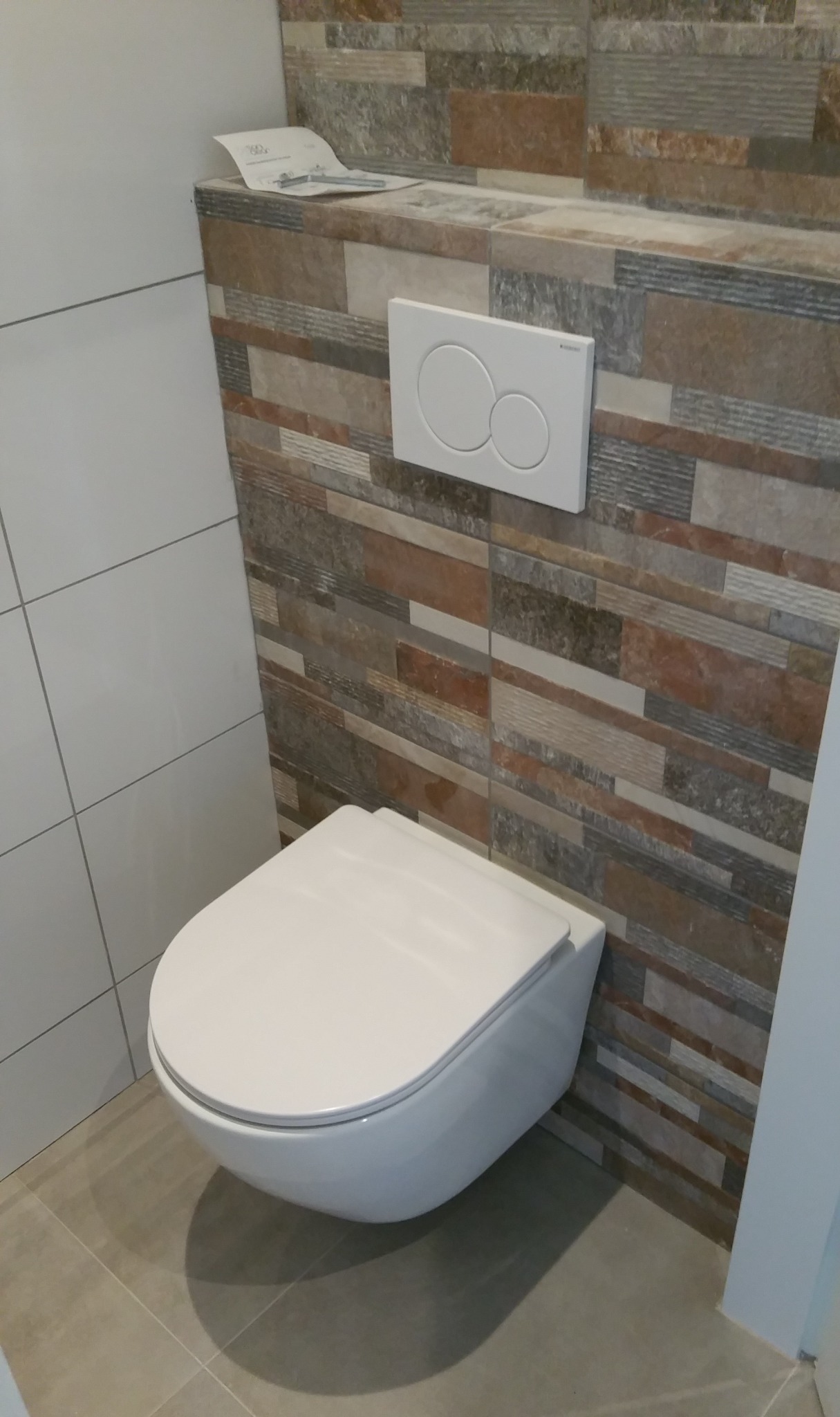 Podwieszana toaleta z białym przyciskiem spłukującym na tle ściany wyłożonej mozaiką z płytek w odcieniach brązu i szarości, z dokumentem na górnej półce.