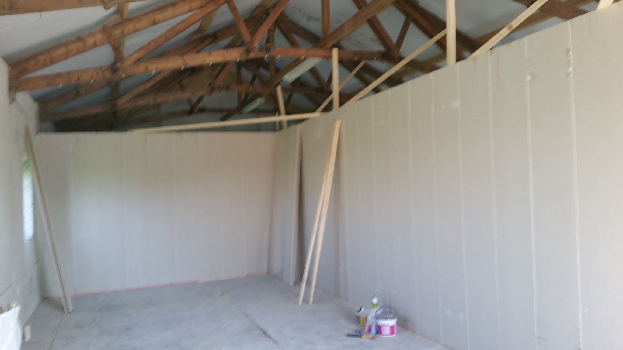 Wnętrze pomieszczenia w trakcie zabudowy płytami gipsowo-kartonowymi, widoczne drewniane elementy konstrukcyjne dachu i ściany z przytwierdzonymi płytami, na podłodze narzędzia i pojemniki z farbą.