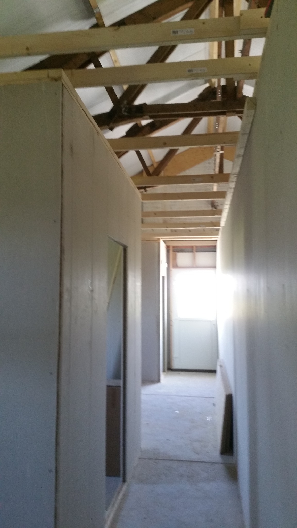 Wnętrze w trakcie budowy: widok korytarza z surowymi ścianami z płyt, drewnianą konstrukcją dachu i jasnym światłem wpadającym z końca korytarza.