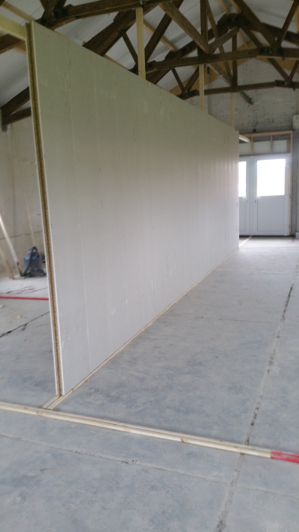 Wnętrze budynku w trakcie prac: wzniesiona ściana z płyt gipsowo-kartonowych na drewnianej konstrukcji szkieletowej, widoczne elementy dachu i podłogi, w tle drzwi.