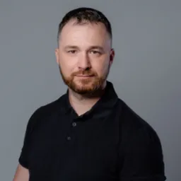 Portret mężczyzny z brodą w ciemnej koszulce polo na szarym tle. Uśmiechnięty, patrzy wprost. Oświetlenie studyjne, równomierne.