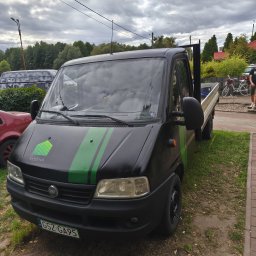 HamBruk - Czarny Fiat Ducato z zielonymi pasami i logo HAMBRUK na masce, zaparkowany na trawie obok chodnika z kostki. Widoczne tablice rejestracyjne i fragment otoczenia.