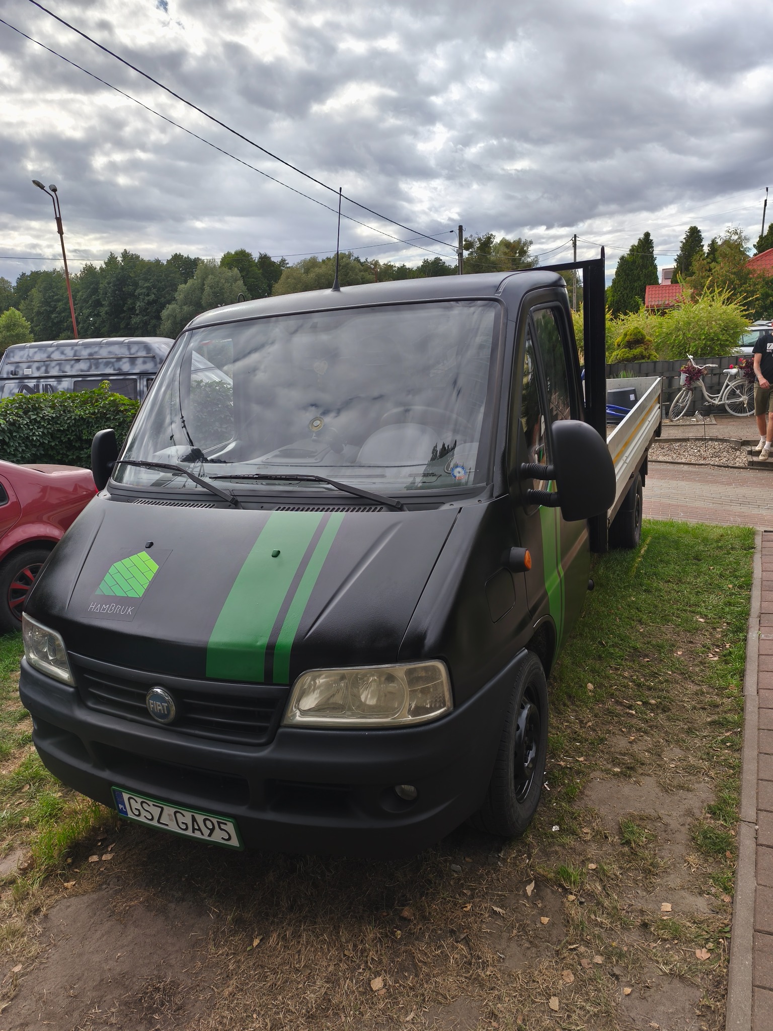Czarny Fiat Ducato z zielonymi pasami i logo HAMBRUK na masce, zaparkowany na trawie obok chodnika z kostki. Widoczne tablice rejestracyjne i fragment otoczenia.