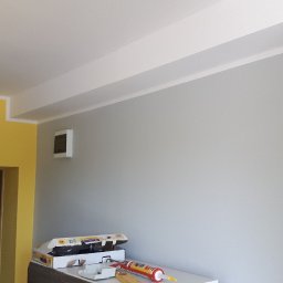 Dariusz Ślesiński - Świeżo pomalowane wnętrze z żółtą i szarą ścianą, widoczne narzędzia malarskie na blacie, sufit z listwami dekoracyjnymi.