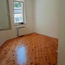 Dariusz Ślesiński - Pomalowane na biało wnętrze pokoju z drewnianą podłogą i grzejnikiem pod oknem. Widoczna lampa wisząca. Świeżo odmalowane ściany.