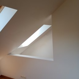 Dariusz Ślesiński - Białe ściany i drewniana podłoga poddasza z oknem dachowym w kształcie trapezu, wnętrze po malowaniu. Widoczne gniazdka elektryczne.