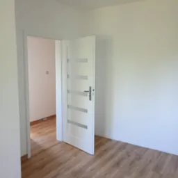 Jasne wnętrze po remoncie: świeżo pomalowane ściany, drewnopodobna podłoga i białe, częściowo otwarte drzwi z przeszkleniami. Widoczne gniazdko elektryczne.