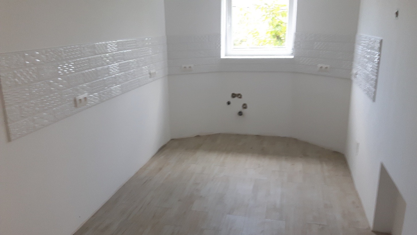 Pusty pokój po remoncie, z nową podłogą imitującą drewno i dekoracyjnymi białymi płytkami na ścianach, widoczne gniazdka elektryczne i wyprowadzone rury, okno z widokiem na zieleń na zewnątrz.