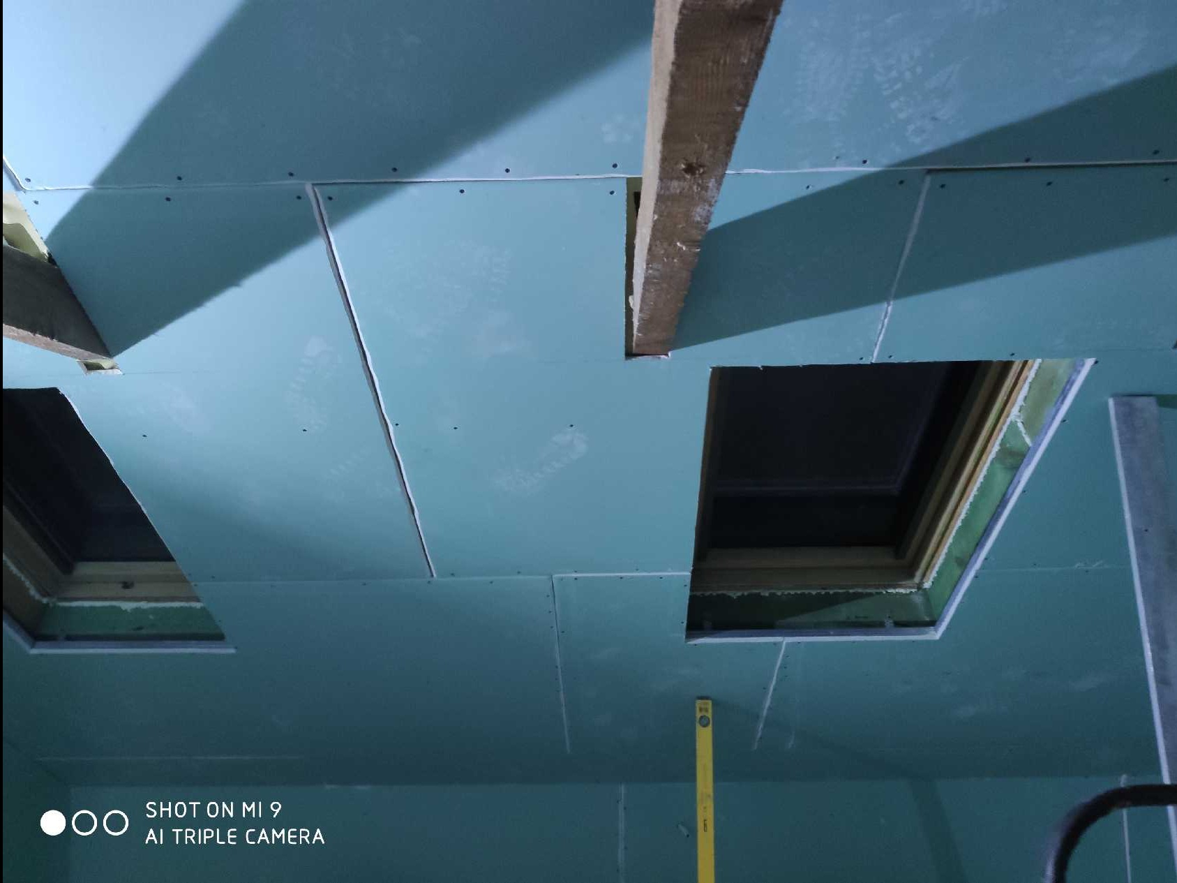 Montaż płyt kartonowo-gipsowych na suficie z widocznymi otworami na okna dachowe i drewnianą belką, poziomica pionowa przy ścianie.