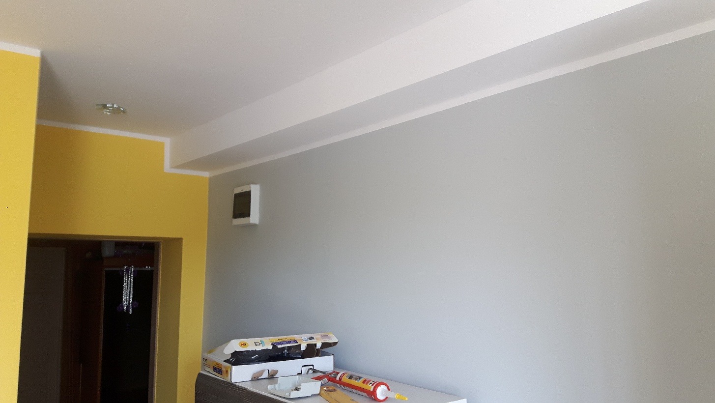 Świeżo pomalowane wnętrze z żółtą i szarą ścianą, widoczne narzędzia malarskie na blacie, sufit z listwami dekoracyjnymi.